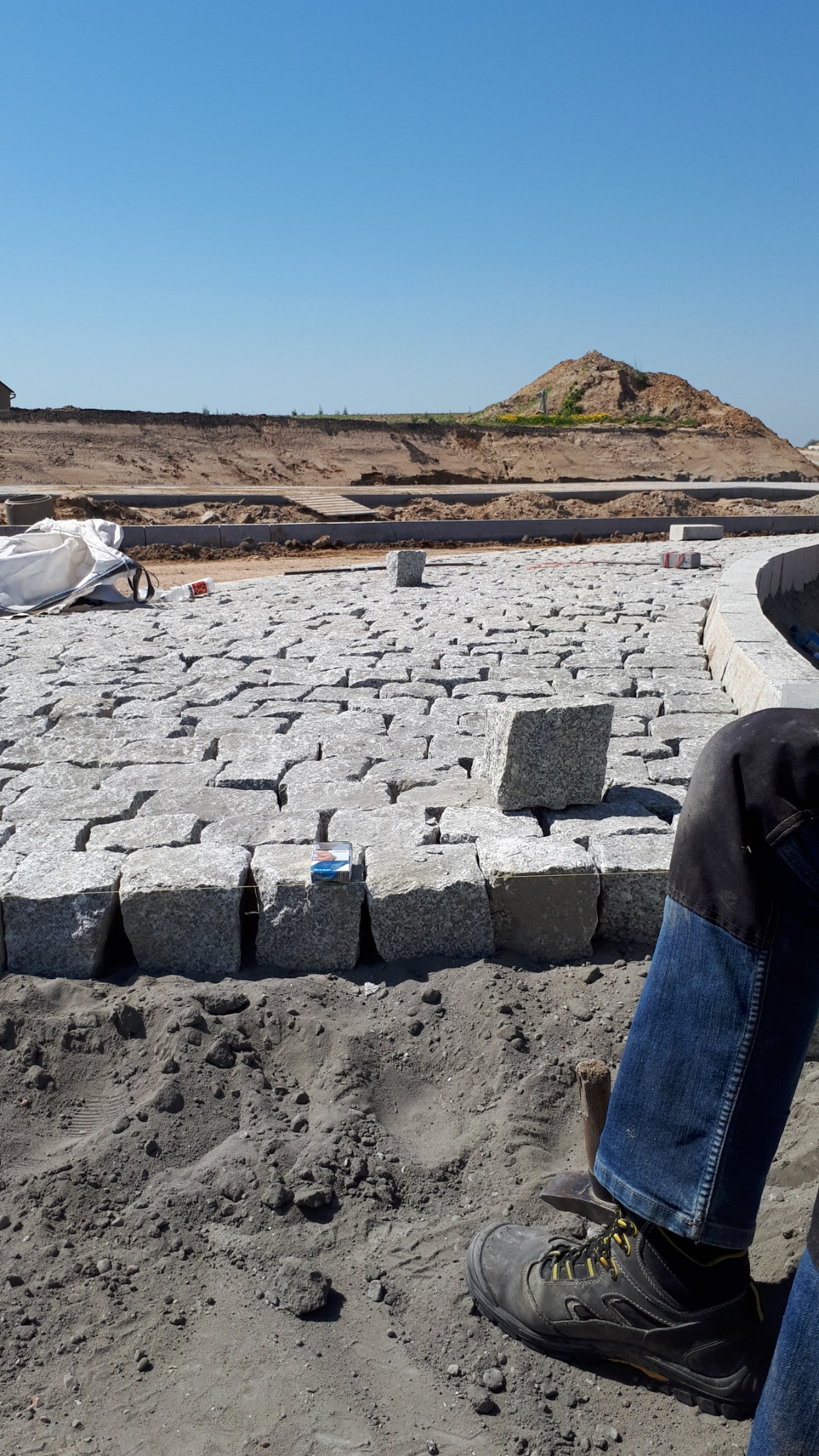 Fragment układanej nawierzchni z kostki granitowej, widoczne niedokończone fragmenty, paczka papierosów położona na kostce, w tle wykop pod budowę i fragment odzieży osoby układającej.