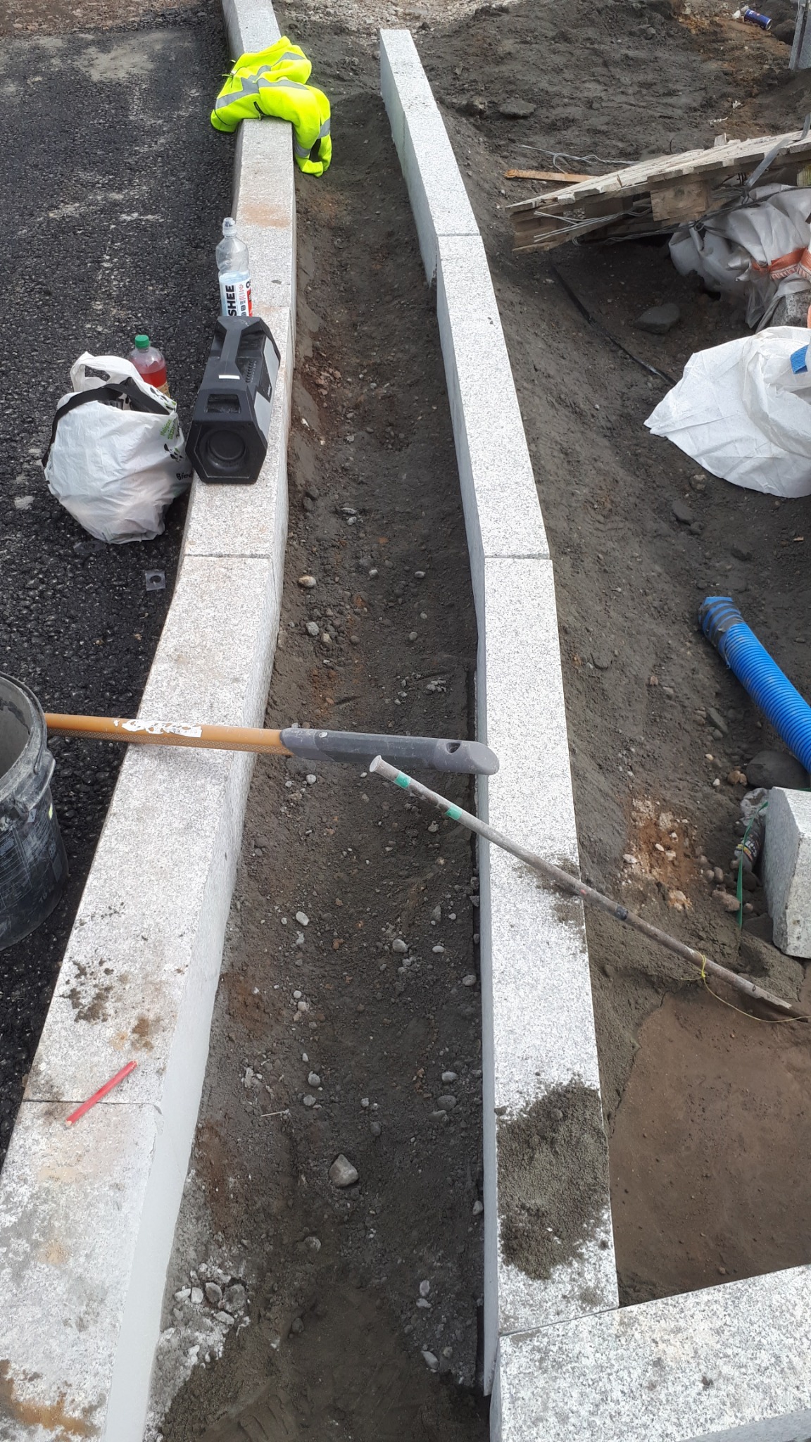 Montaż obrzeży z jasnoszarego granitu, widoczne narzędzia, odzież robocza i głośnik przenośny.