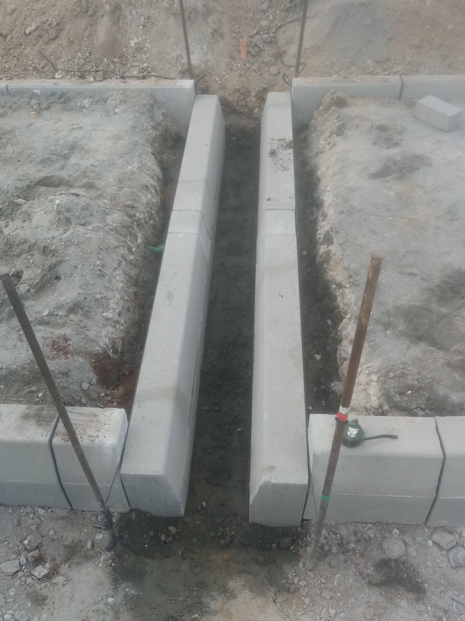 Przygotowanie podłoża pod kostkę brukową z ustawionymi krawężnikami i wbitymi palikami, widoczna ziemia i częściowo ułożona kostka brzegowa.