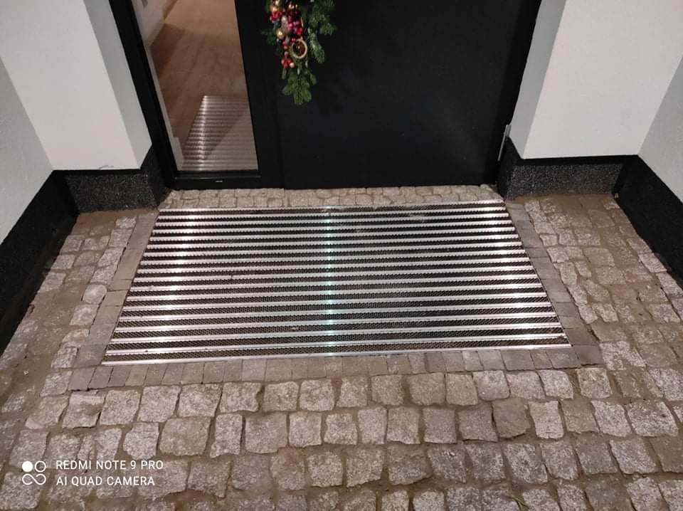 Wejście do domu z kostki granitowej, wykończone metalową wycieraczką z poziomymi paskami, ozdobione świątecznym wieńcem na czarnych drzwiach.