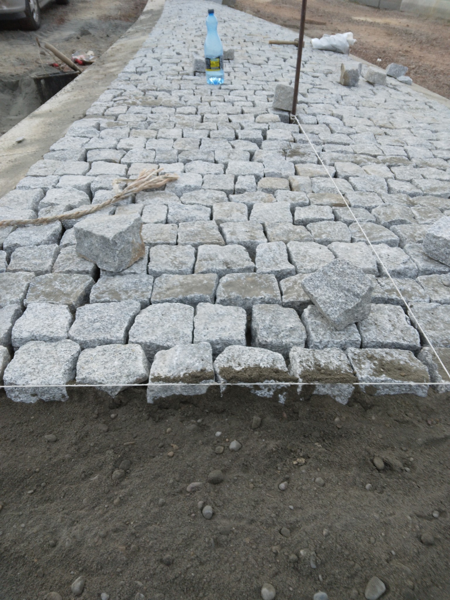 Układanie szarej kostki granitowej na podsypce piaskowej, widoczny sznurek wyznaczający linię, butelka wody i narzędzia w tle.