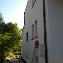 Świeżo otynkowana ściana domu jednorodzinnego z widoczną drabiną, narzędziami i skrzynką elektryczną, z czarną rynną spustową i oknami, częściowo widoczny teren budowy z drewnem.