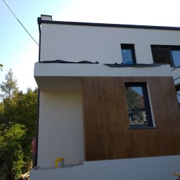 TOPWORKER SP. Z O.O. - Fragment nowo wybudowanego domu z jasną elewacją i drewnianą okładziną, widoczne okna z czarnymi ramami i elementy wykończeniowe, w tle zieleń drzew i niebo.