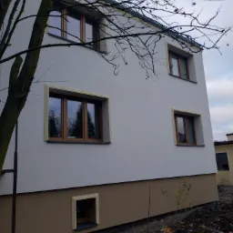 Dwukondygnacyjny dom z oknami w drewnianych ramach i kremową elewacją, z dolną częścią w kolorze brązowym. Widoczne drzewo z nagimi gałęziami na tle pochmurnego nieba.