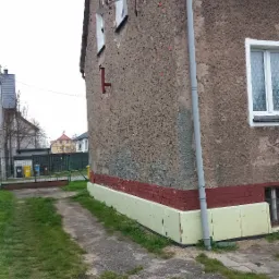 Fragment ściany starego domu z widoczną warstwą izolacyjną fundamentów w postaci jasnych płyt, częściowo pomalowaną na kolor czerwony, z widoczną starą elewacją i rynną spustową.