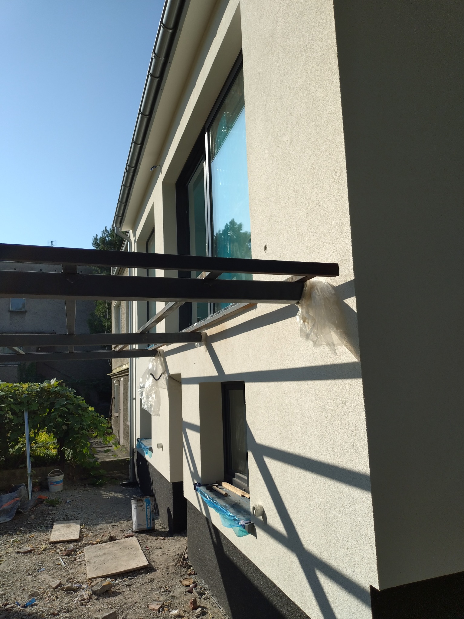 Świeżo otynkowana elewacja domu jednorodzinnego z zamontowanymi oknami, widoczne metalowe elementy konstrukcyjne z folią ochronną i otoczenie placu budowy z elementami materiałów budowlanych.