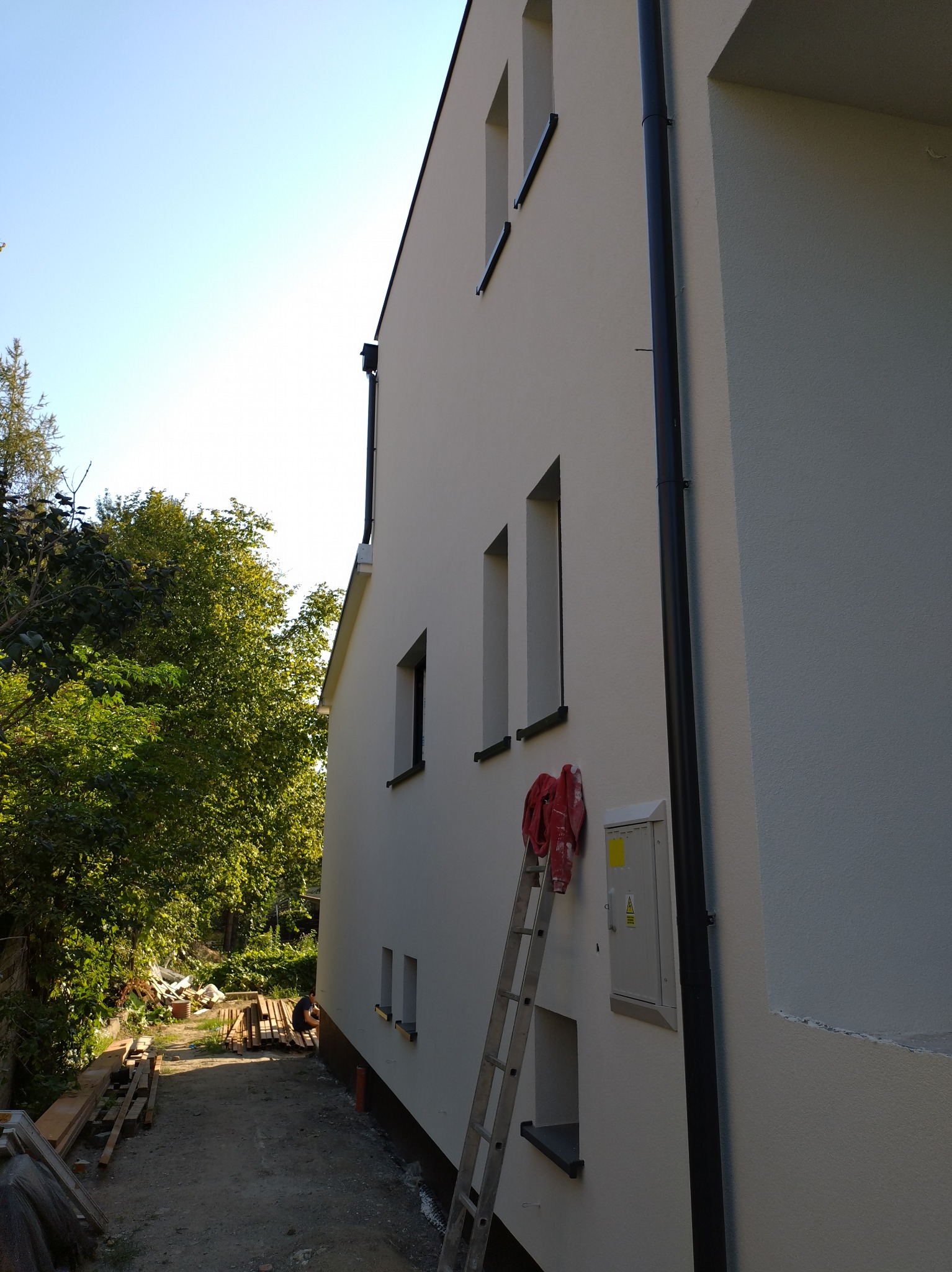 Świeżo otynkowana ściana domu jednorodzinnego z widoczną drabiną, narzędziami i skrzynką elektryczną, z czarną rynną spustową i oknami, częściowo widoczny teren budowy z drewnem.