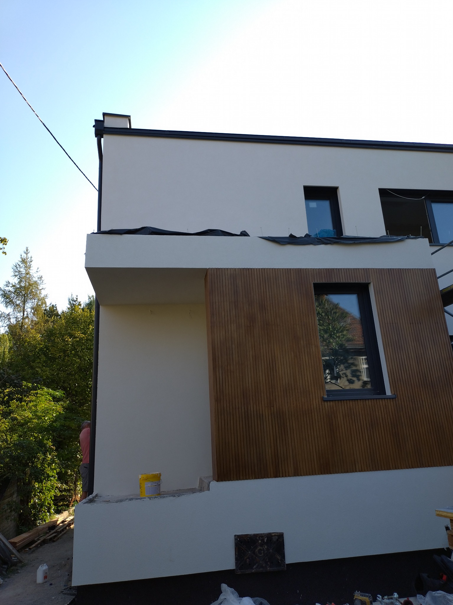 Fragment nowo wybudowanego domu z jasną elewacją i drewnianą okładziną, widoczne okna z czarnymi ramami i elementy wykończeniowe, w tle zieleń drzew i niebo.