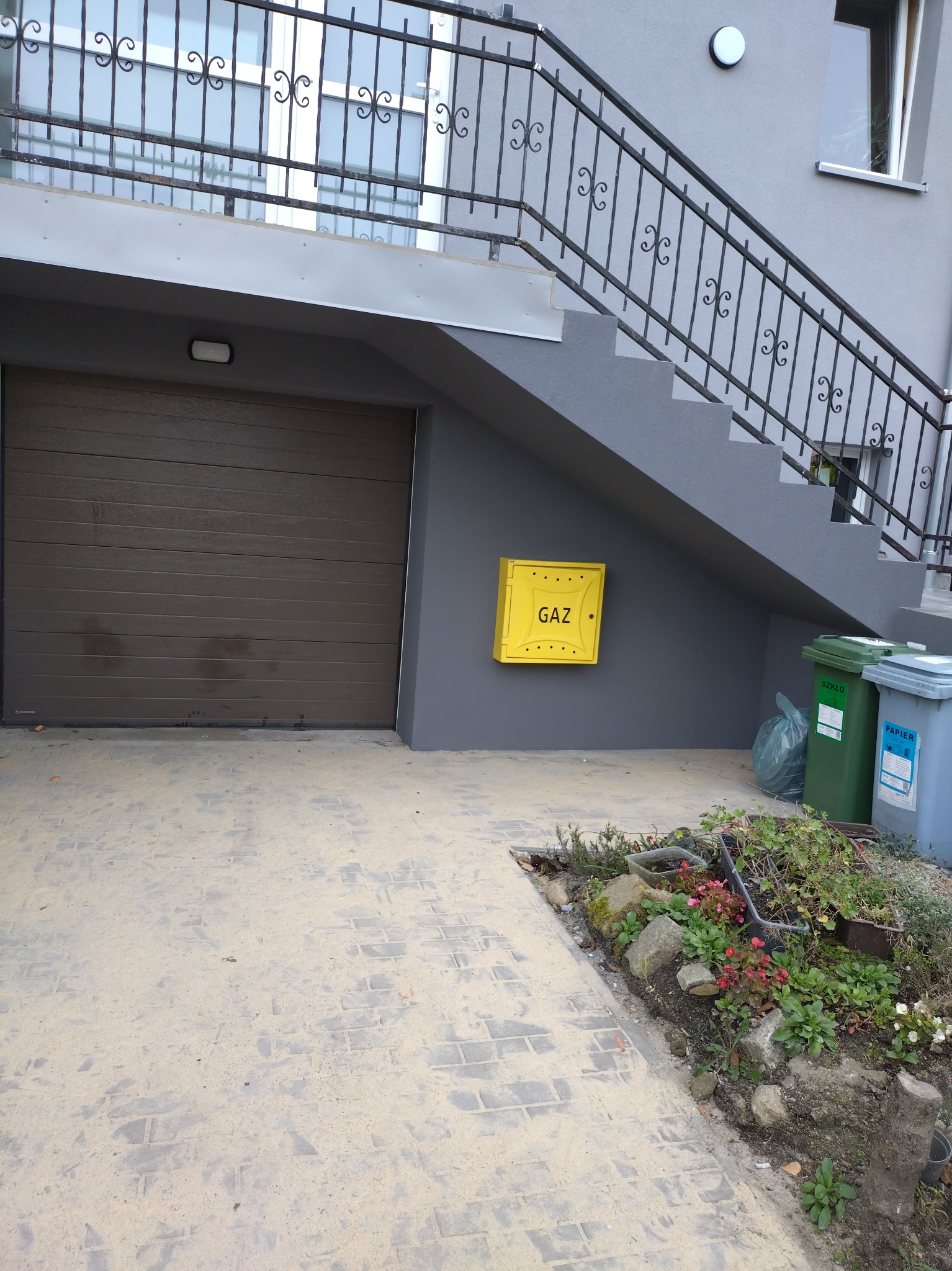 Szary dom z brązową bramą garażową, schodami z kutą balustradą, żółtą skrzynką gazową i dwoma pojemnikami na śmieci obok rabaty kwiatowej.