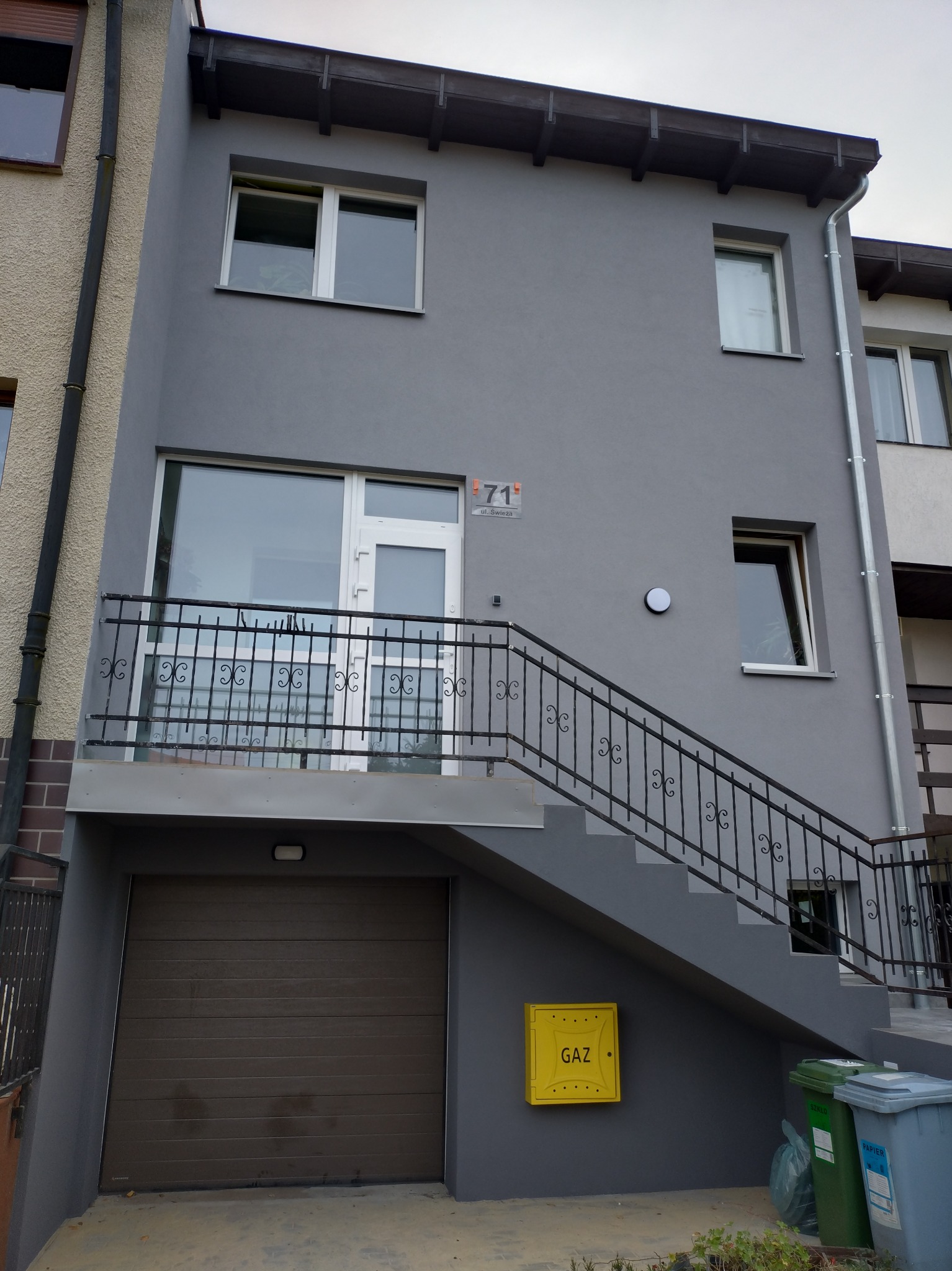 Dwukondygnacyjny dom z garażem i zewnętrznymi schodami, pomalowany na szaro, z żółtą skrzynką gazową i plastikowymi pojemnikami na śmieci przy schodach.