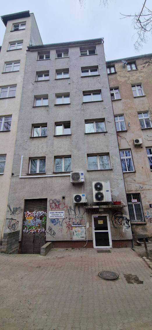 Szary budynek wielorodzinny z widocznymi śladami zabrudzeń i graffiti na elewacji parteru, z trzema klimatyzatorami na ścianie i dwoma wejściami.