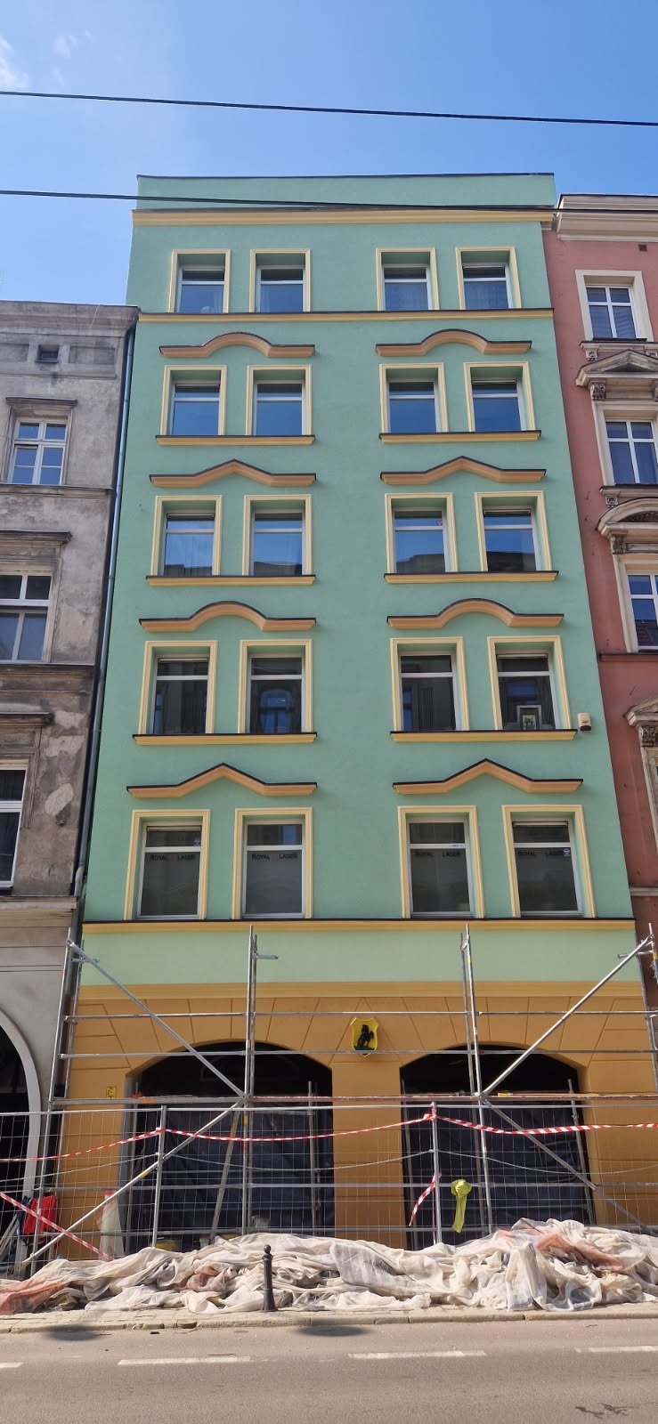 Odnowiona, pastelowa elewacja kamienicy z żółtymi akcentami, częściowo zasłonięta rusztowaniem. Widoczne okna z refleksami światła i sąsiednie budynki.