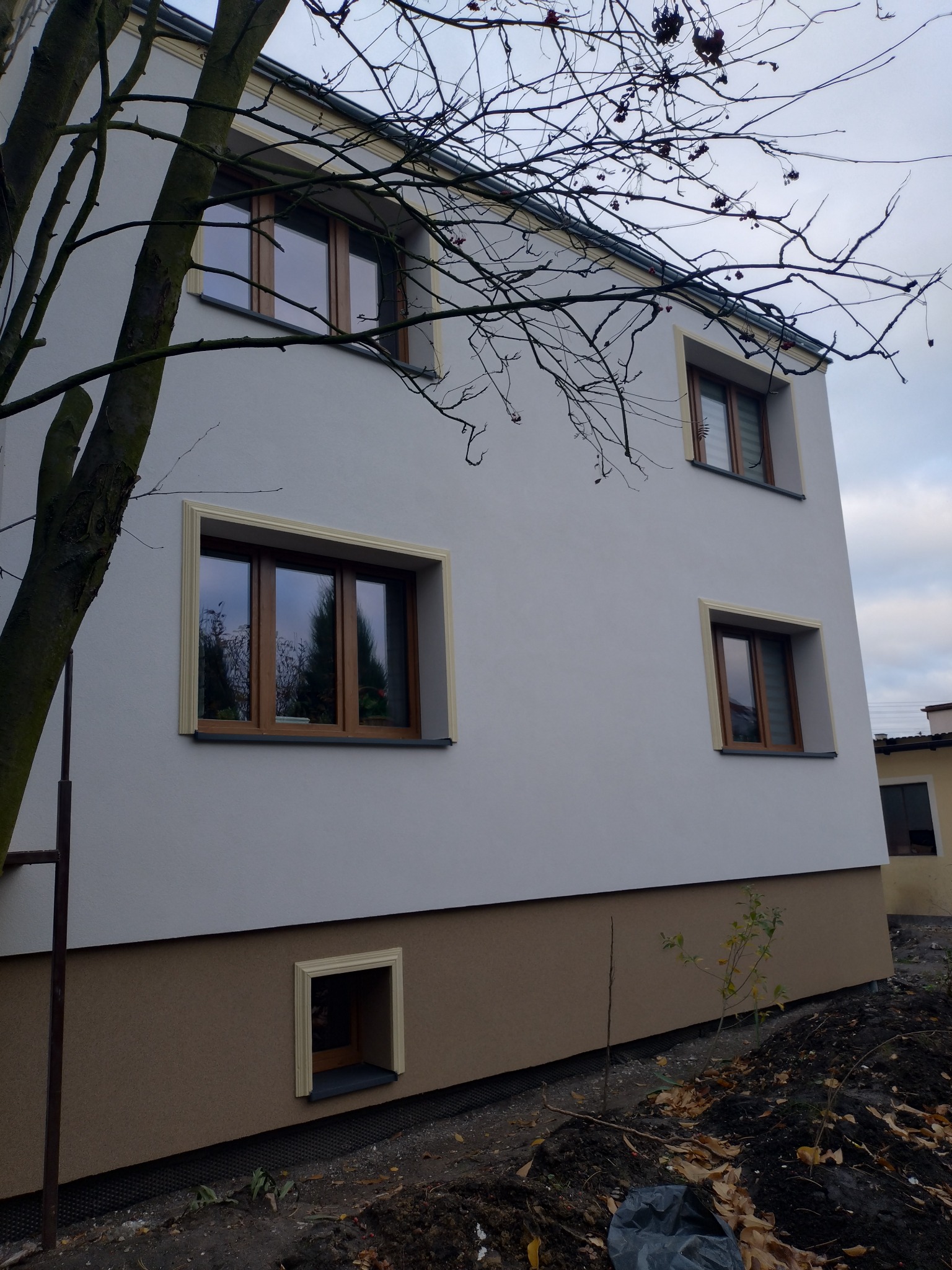 Dwukondygnacyjny dom z oknami w drewnianych ramach i kremową elewacją, z dolną częścią w kolorze brązowym. Widoczne drzewo z nagimi gałęziami na tle pochmurnego nieba.