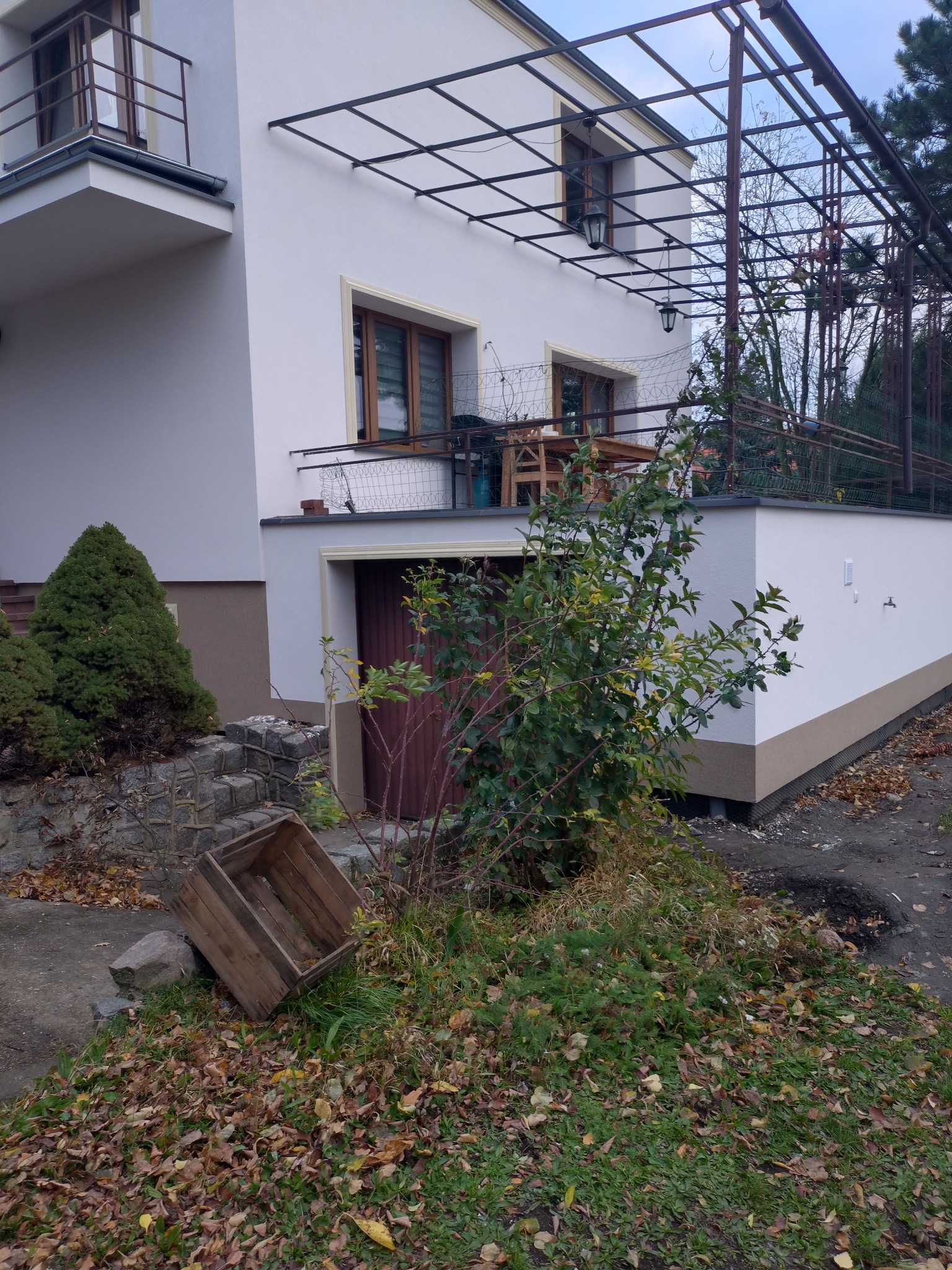 Dwupiętrowy dom z białymi ścianami i brązowymi akcentami, widoczny z poziomu gruntu, z drewnianą skrzynią na pierwszym planie i metalową konstrukcją nad balkonem.