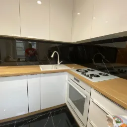 Nowoczesna kuchnia z białymi, lakierowanymi frontami, blatem w kolorze jasnego drewna i czarnym panelem szklanym nad blatem z białymi żyłami, odbijającym sylwetkę osoby w oknie.