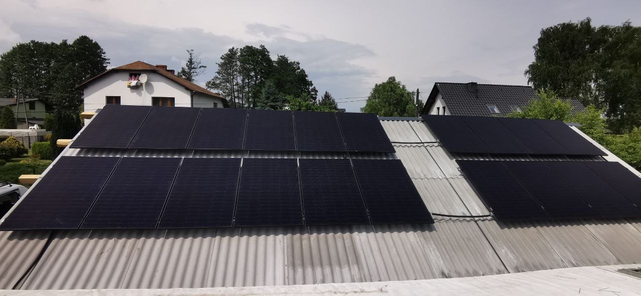 Panele słoneczne zamontowane na pofałdowanym, szarym dachu budynku mieszkalnego, widok z poziomu dachu, z domami i drzewami w tle.