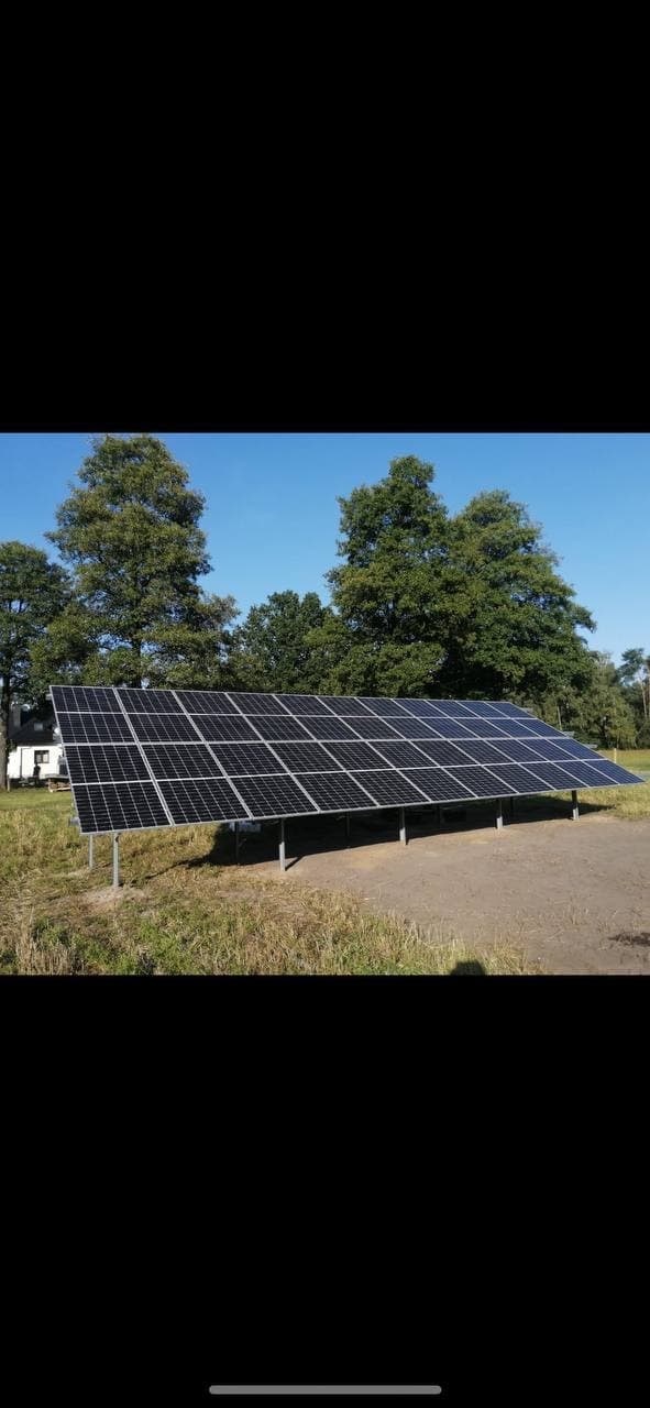 Panel słoneczny na metalowej konstrukcji ustawiony na trawiastym terenie, z drzewami i błękitnym niebem w tle.