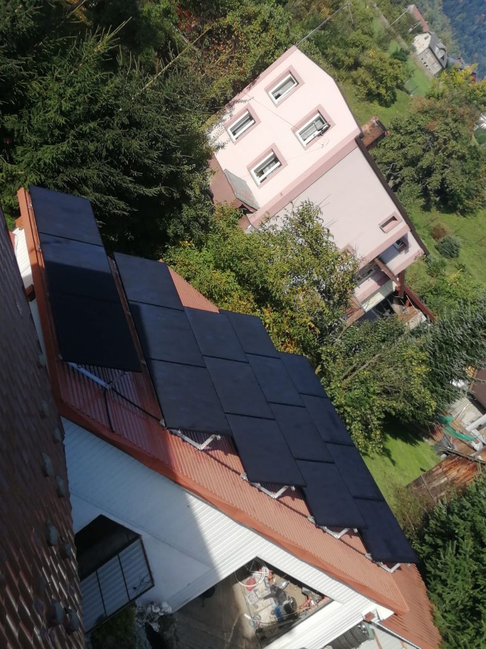 Panele słoneczne zamontowane na dachu domu z brązową dachówką, widoczne z góry, w tle budynek mieszkalny i drzewa.