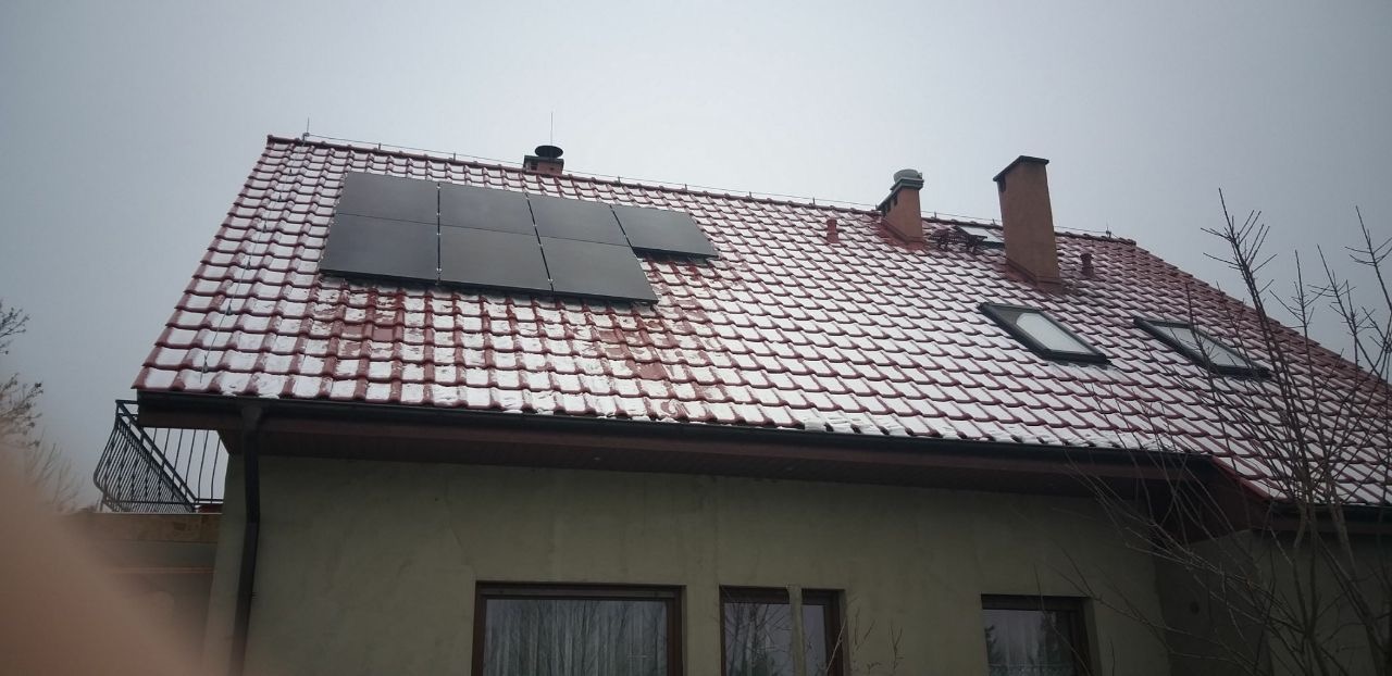 Częściowo ośnieżony dach domu z zamontowanymi panelami słonecznymi; widoczne kominy i okna dachowe.