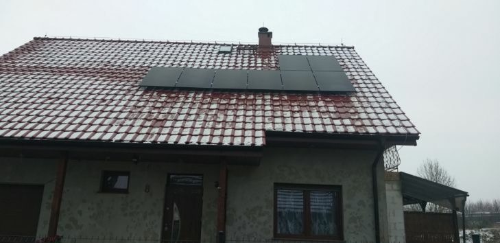 Panele słoneczne na dachu domu pokrytym śniegiem i czerwoną dachówką, widok z poziomu gruntu w pochmurny dzień.