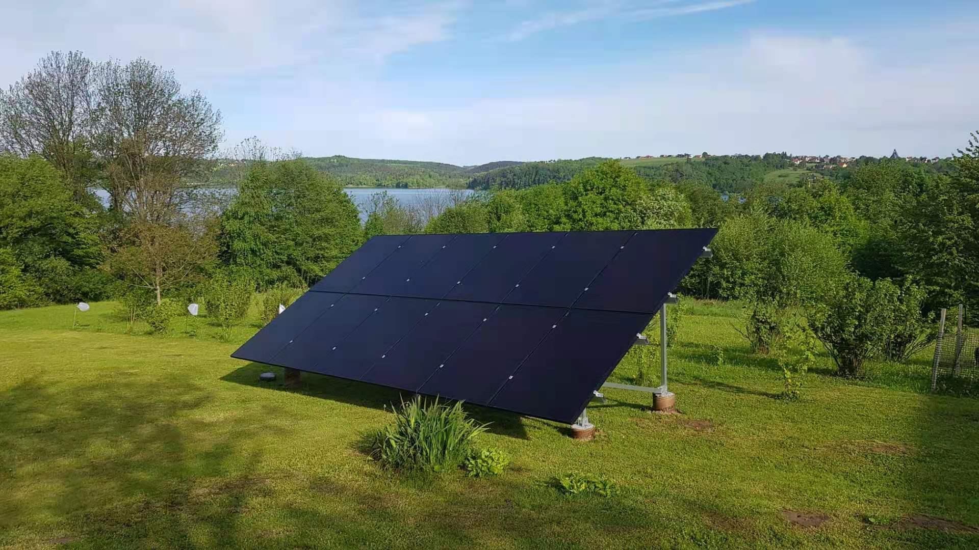 Czarny panel fotowoltaiczny ustawiony na metalowej konstrukcji na trawiastym wzgórzu z widokiem na jezioro i las w tle.