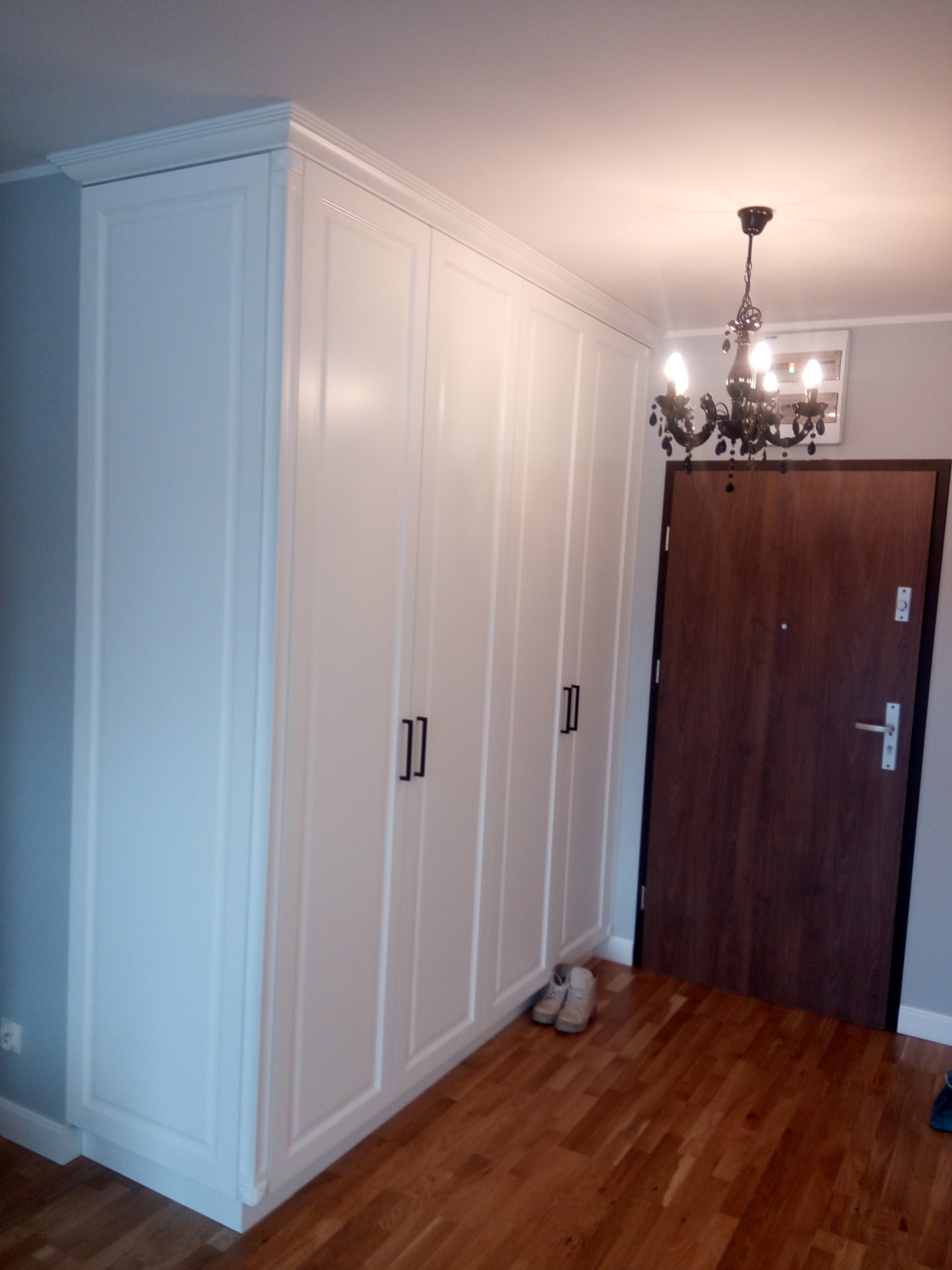 Biała, wysoka szafa z czterema drzwiami i czarnymi uchwytami, sięgająca sufitu, z dekoracyjnym gzymsem, na tle drewnianej podłogi i brązowych drzwi wejściowych, oświetlona żyrandolem.