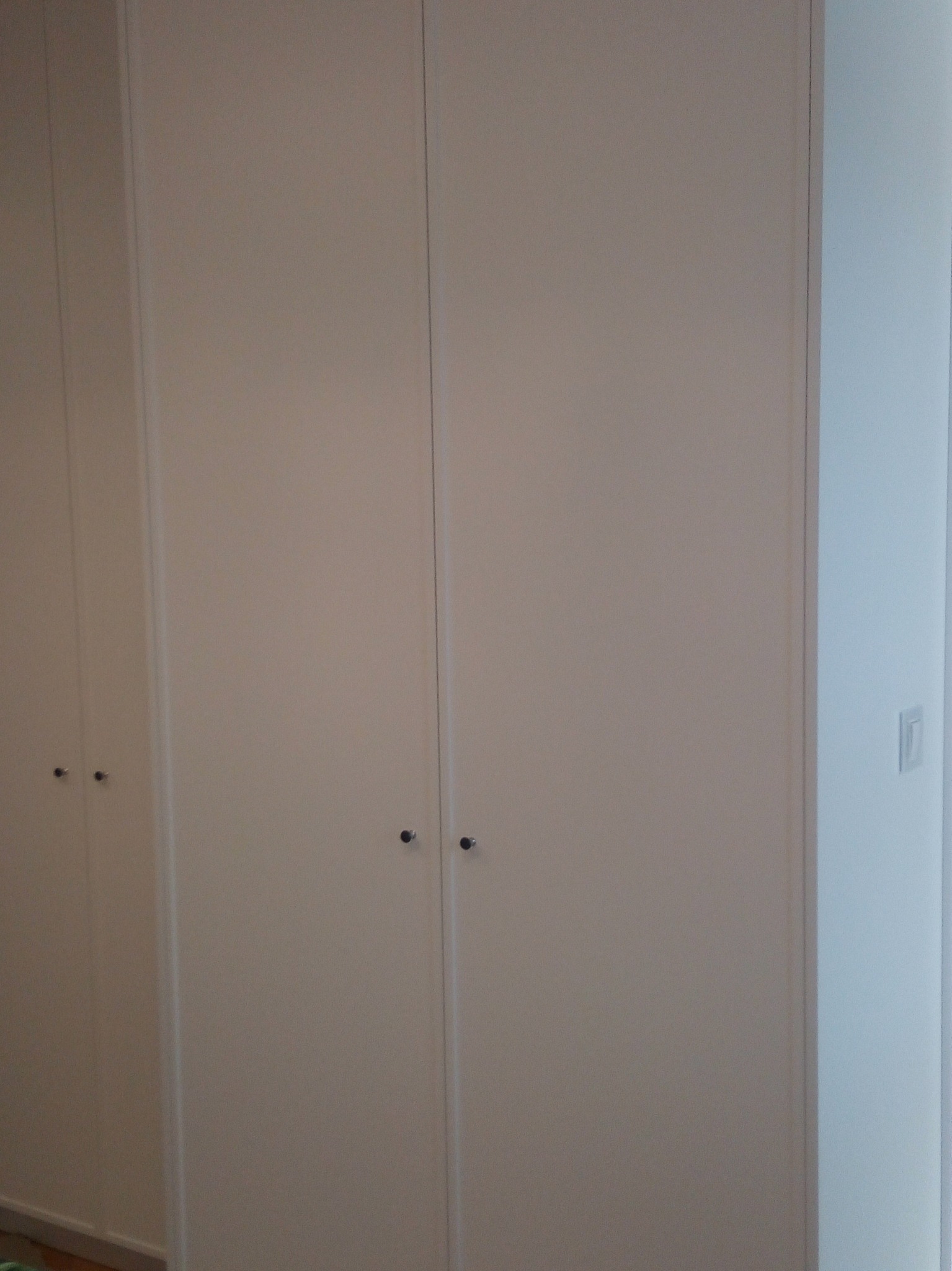 Zamknięta, biała szafa wnękowa z małymi, okrągłymi uchwytami w Dębem Wielkim, widoczna z bliskiej perspektywy, z fragmentem ściany po prawej stronie.