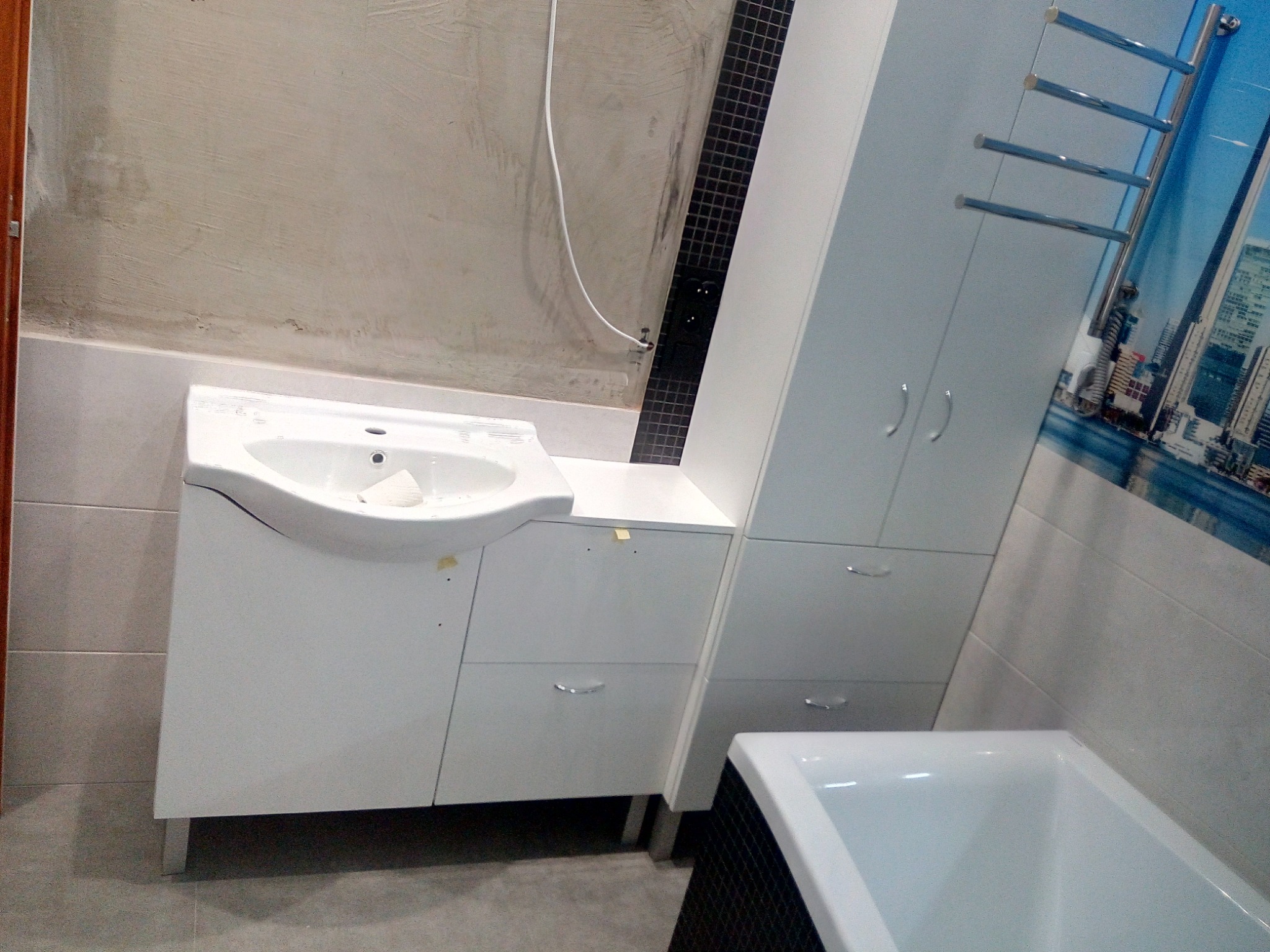 Niedokończona łazienka z białą szafką pod umywalką, wysoką szafą z grzejnikiem drabinkowym i częściowo obłożoną wanną, widoczne ślady po pracach wykończeniowych na ścianach.