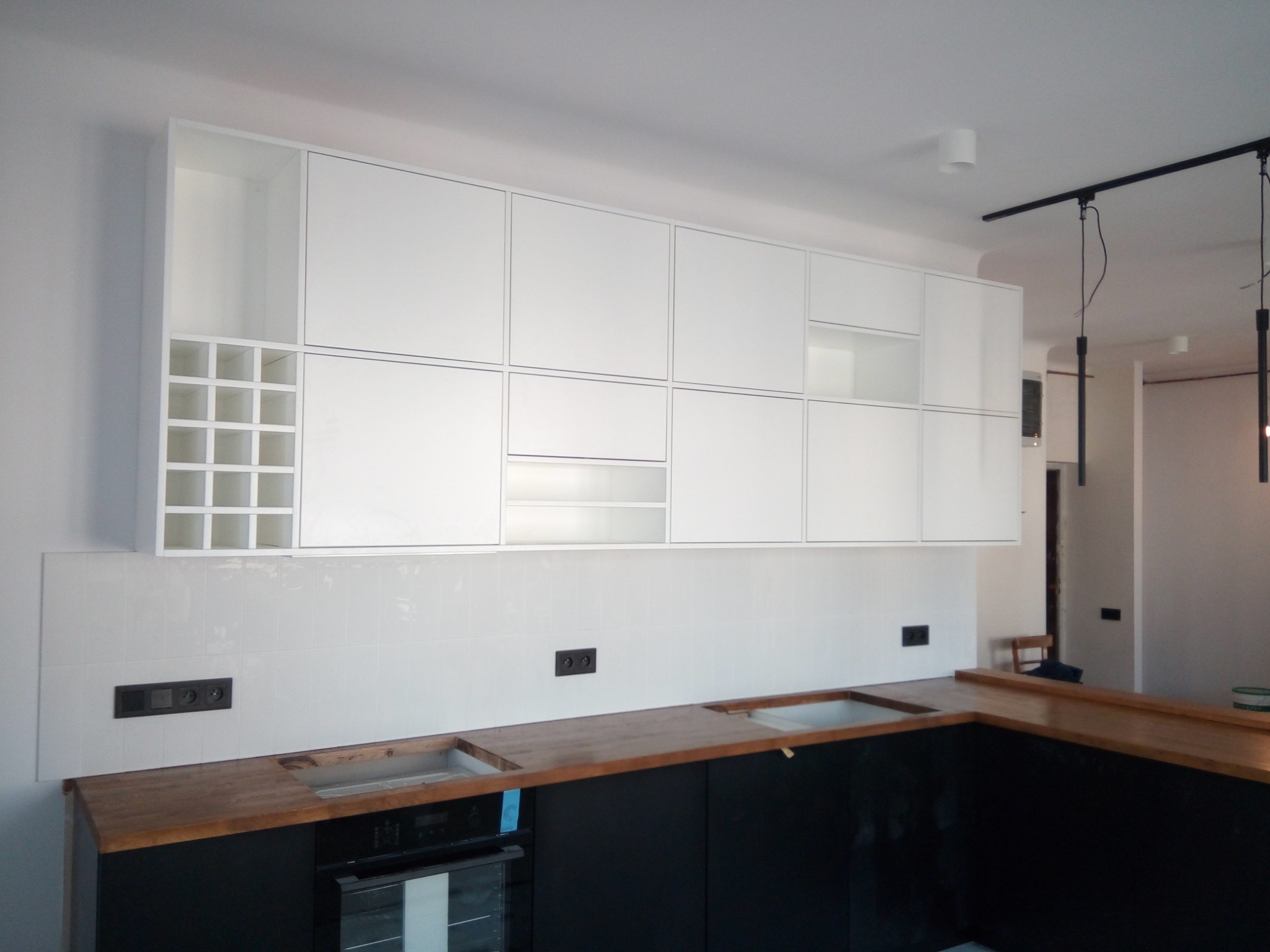 Białe szafki kuchenne wiszące z modułem na wino, czarne szafki dolne z drewnianym blatem w trakcie montażu, widoczne gniazdka elektryczne i brakujące zlewozmywaki.