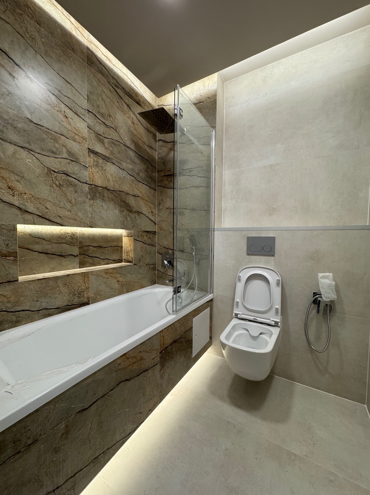 Wykończona łazienka w Wołominie: marmurowe płytki, podświetlana wnęka nad wanną, szklany parawan nawannowy i podwieszana toaleta. Nowoczesny design.
