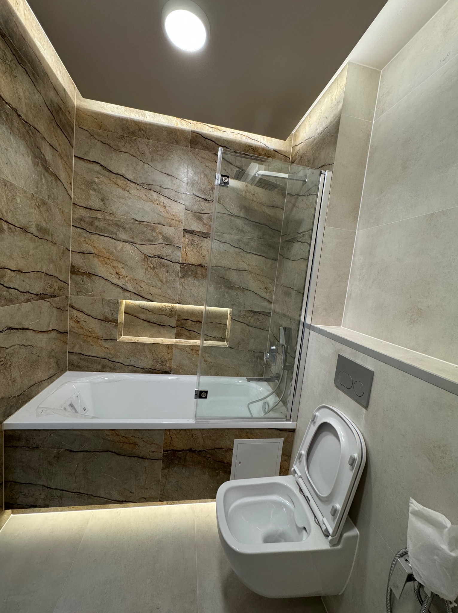 Nowoczesna łazienka z marmurowymi płytkami, podświetlaną wnęką i kabiną prysznicową. Uwagę zwraca wisząca toaleta z otwartą klapą i minimalistyczny design.