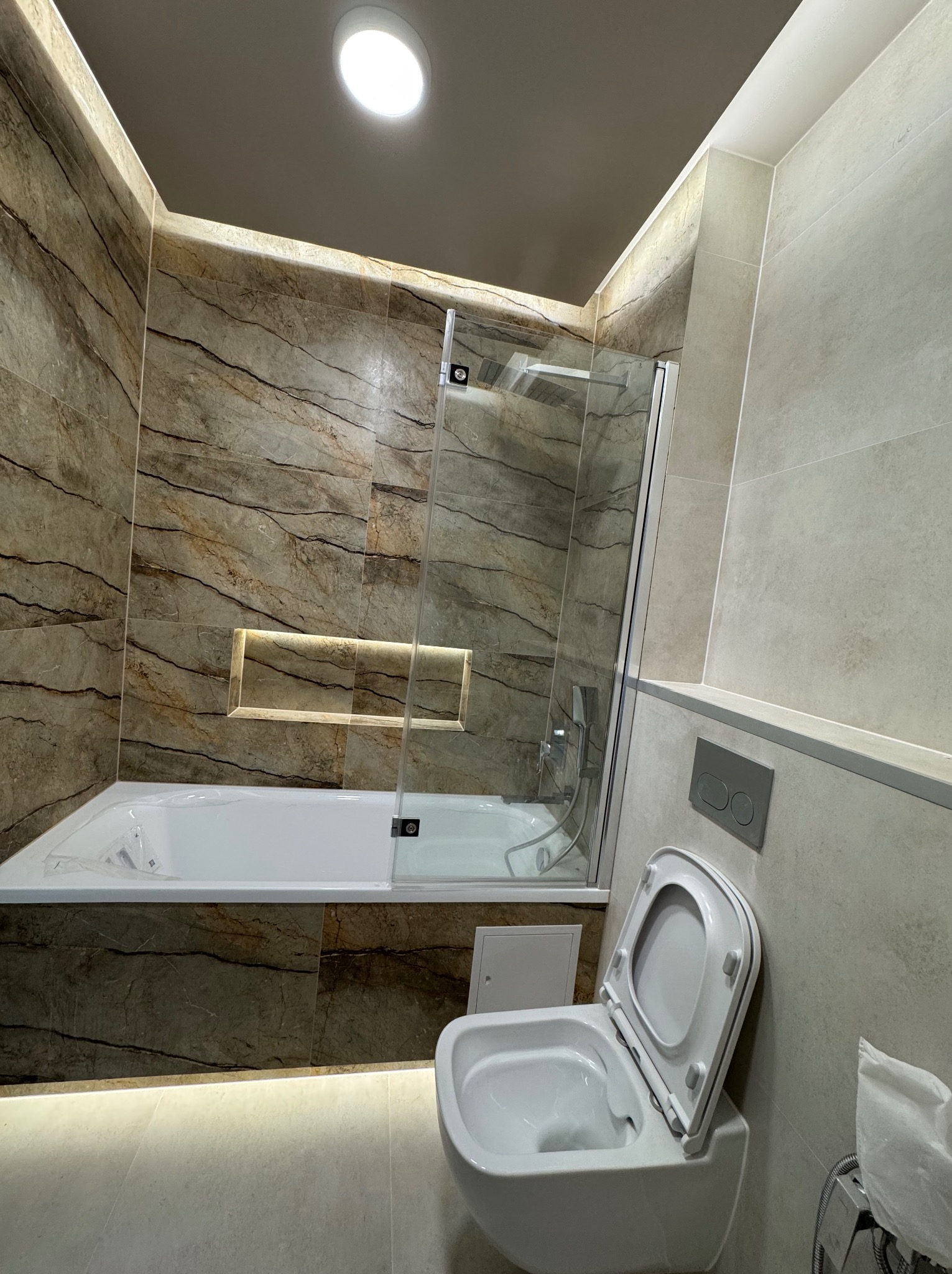 Wykończona łazienka z marmurowymi płytkami, podświetlaną wnęką i nowoczesną toaletą podwieszaną. Kabina prysznicowa ze szklanym panelem.