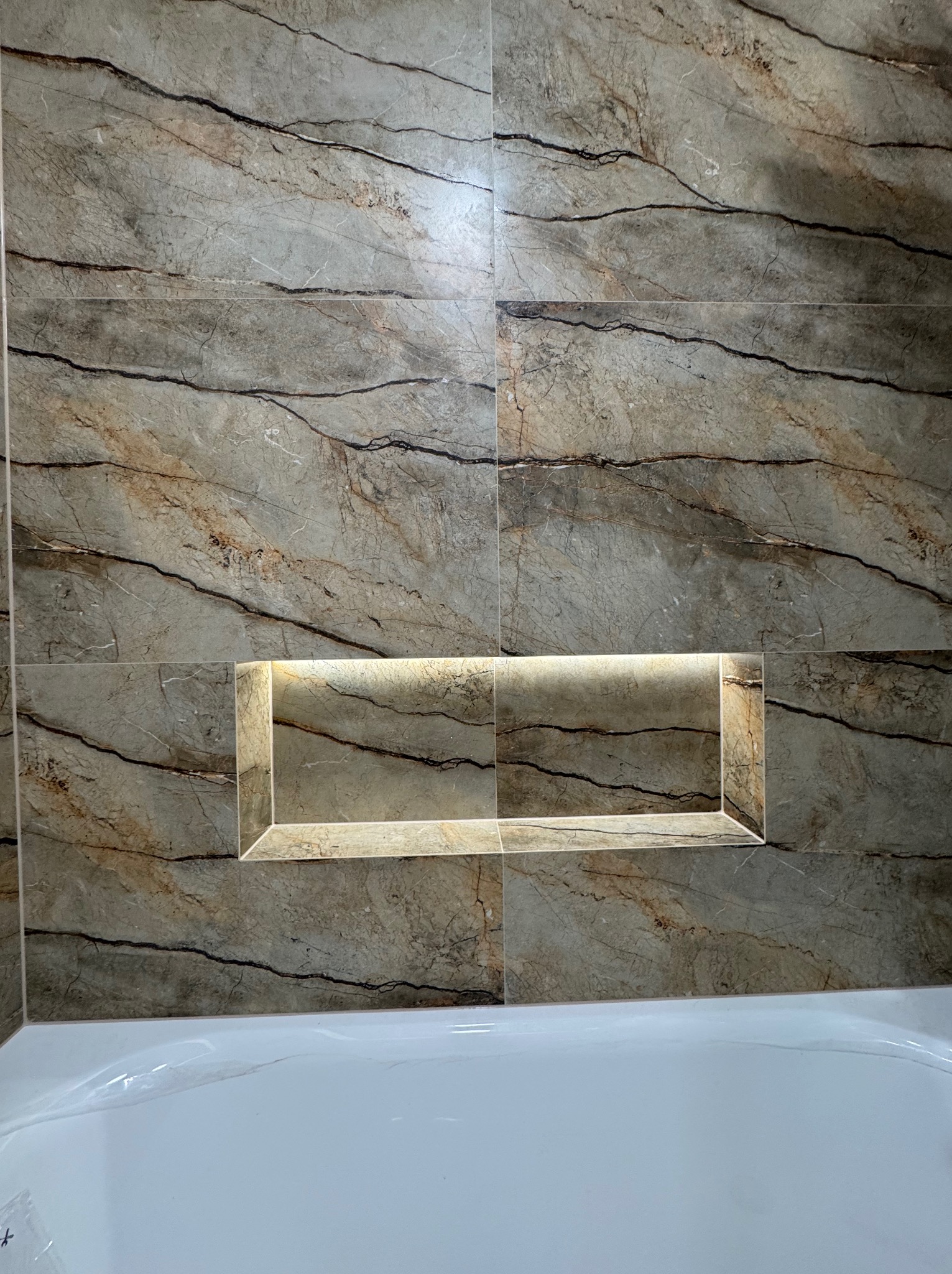 Wykończona ściana nad wanną z płytek imitujących marmur, z podświetlaną wnęką. Estetyczne połączenie materiałów i oświetlenia w łazience.