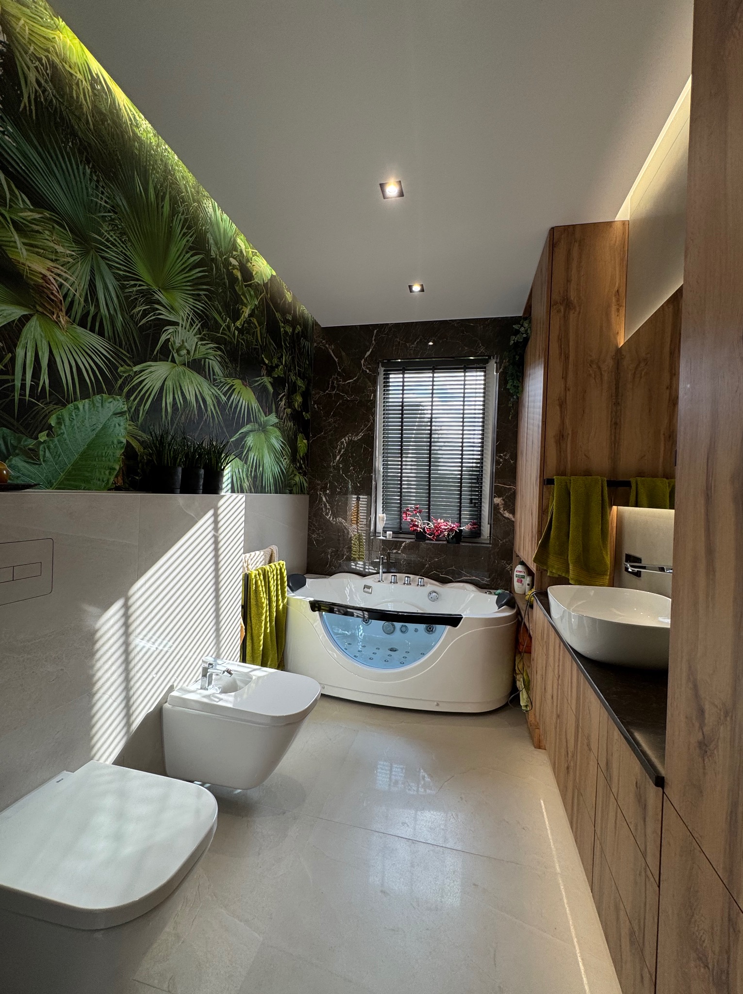 Nowoczesna łazienka z wanną jacuzzi, bidetem i toaletą w jasnych barwach. Na ścianie fototapeta z motywem tropikalnych liści. Drewniane szafki i marmurowe akcenty.
