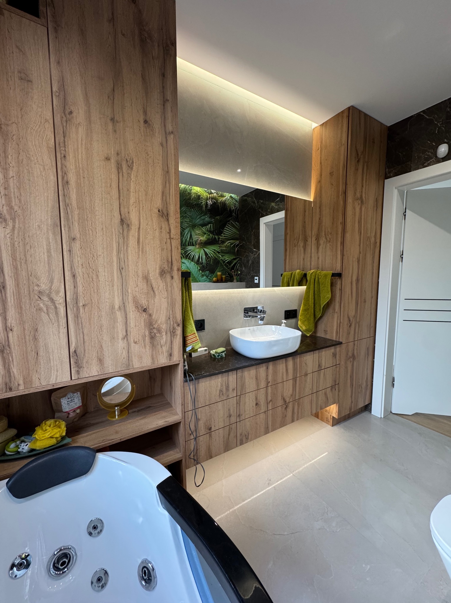 Nowoczesna łazienka z drewnianymi szafkami, owalną umywalką i lustrem z podświetleniem LED. W tle widoczna wanna z hydromasażem i roślinny motyw na ścianie.