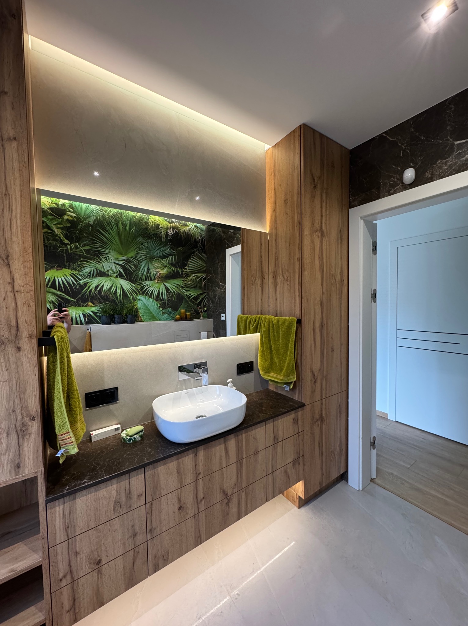Nowoczesna łazienka z drewnianą zabudową, umywalką nablatową i lustrem z motywem roślinnym. Oświetlenie LED podkreśla elegancję wnętrza. Widoczny fragment otwartych drzwi.