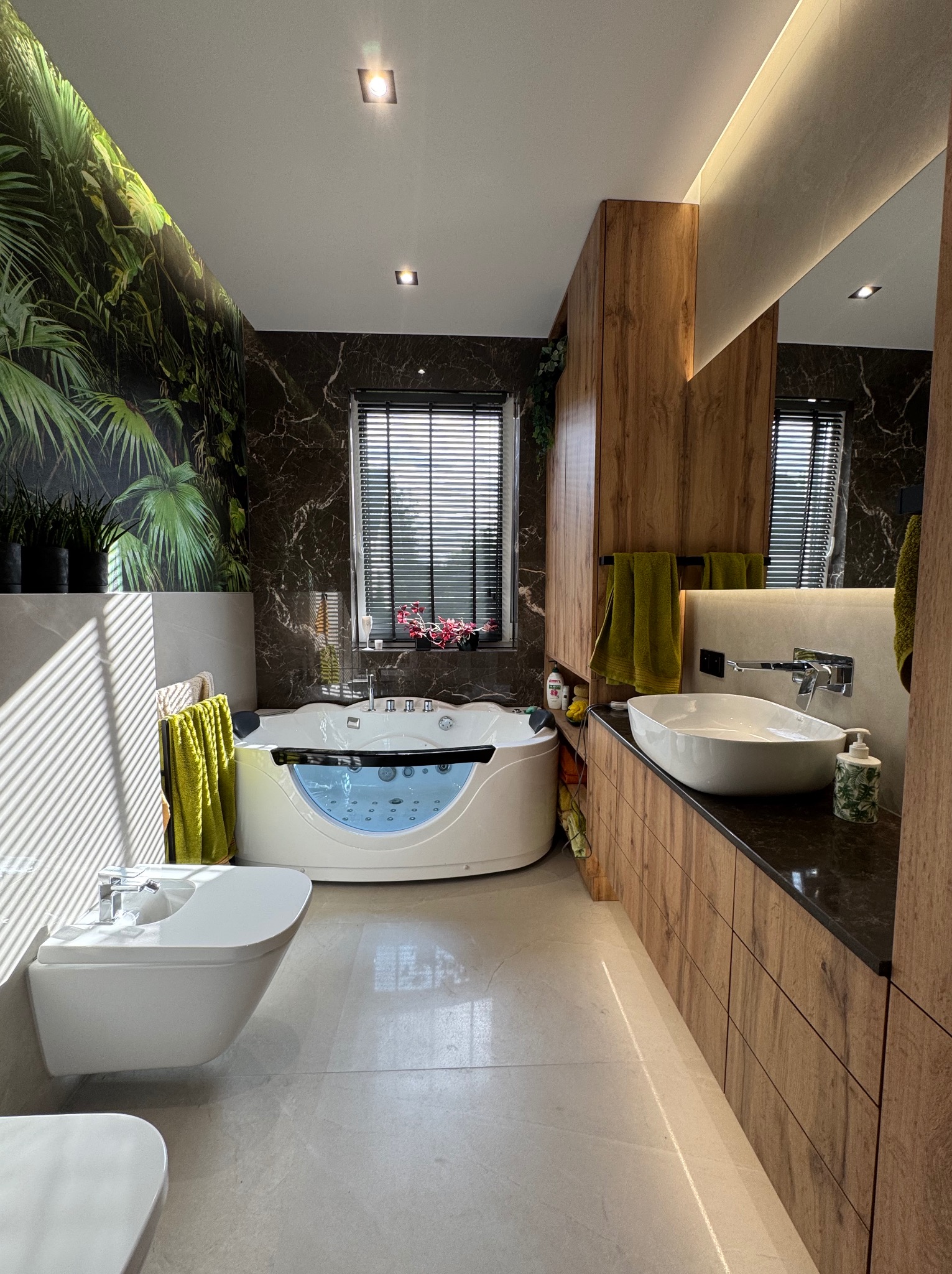 Nowoczesna łazienka z wanną z hydromasażem, bidetem i umywalką nablatową. Ciemne marmurowe ściany kontrastują z drewnianymi szafkami i zielonym akcentem roślinnym.