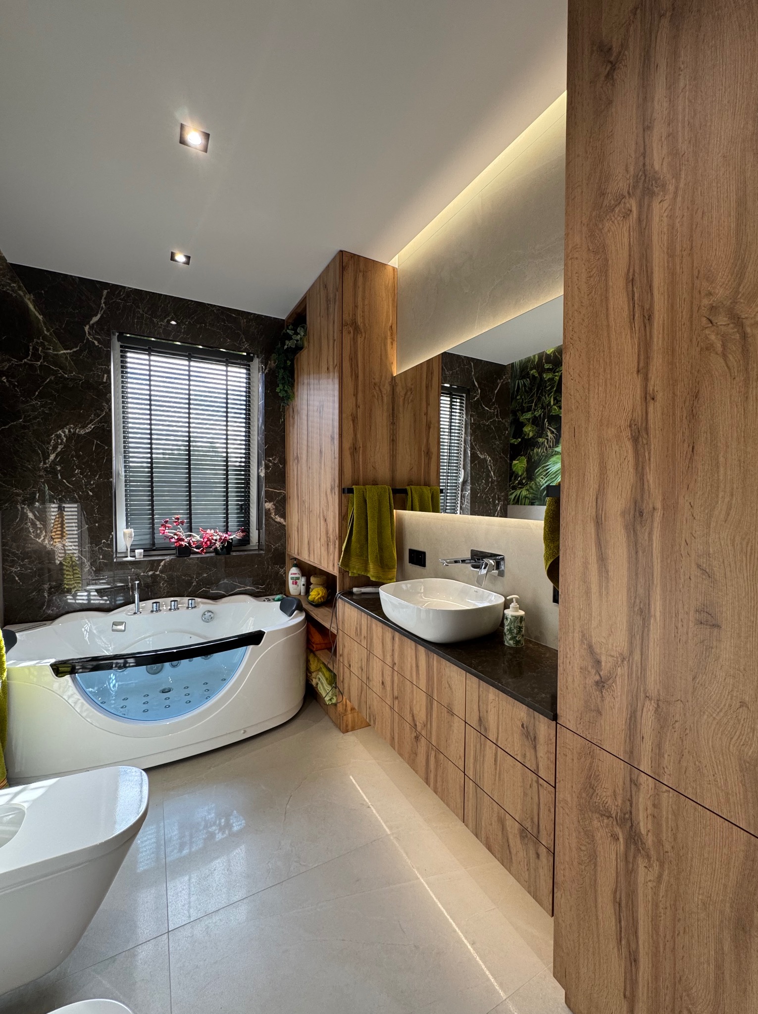 Nowoczesna łazienka z wanną z hydromasażem, umywalką nablatową i drewnianymi szafkami. Marmurowe ściany i rośliny dodają elegancji. Oświetlenie LED podkreśla detale.