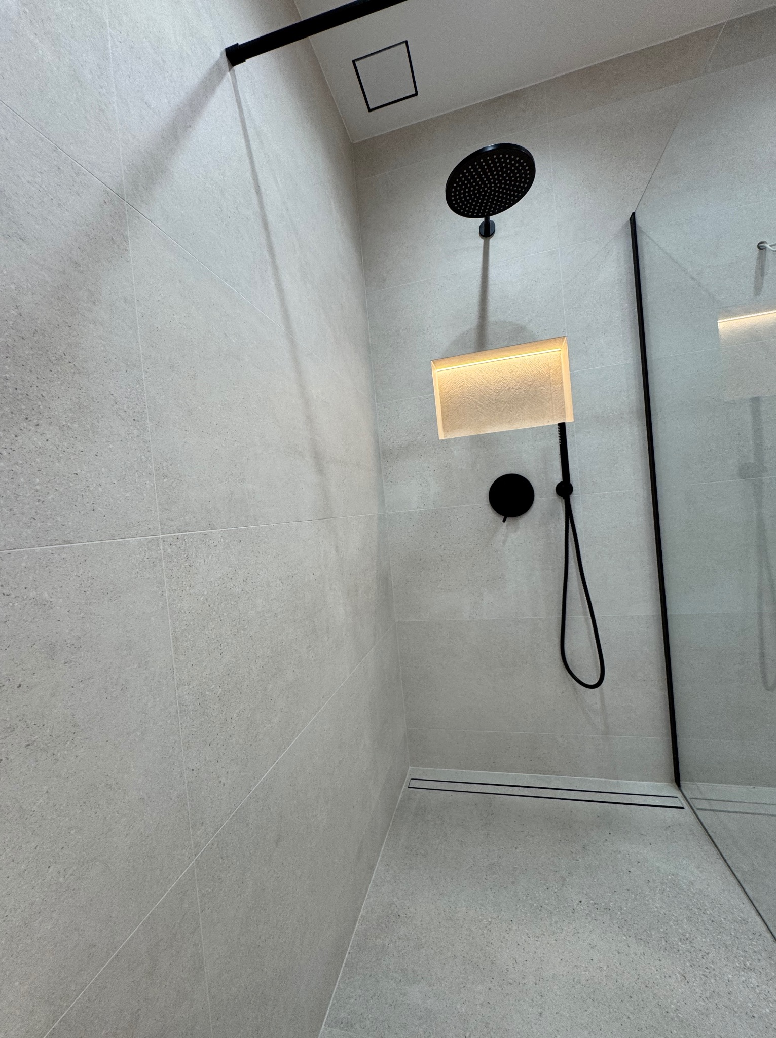 Nowoczesna kabina prysznicowa z szarymi płytkami, czarną armaturą i podświetlaną wnęką. Minimalistyczny design z odpływem liniowym i szklaną ścianką.