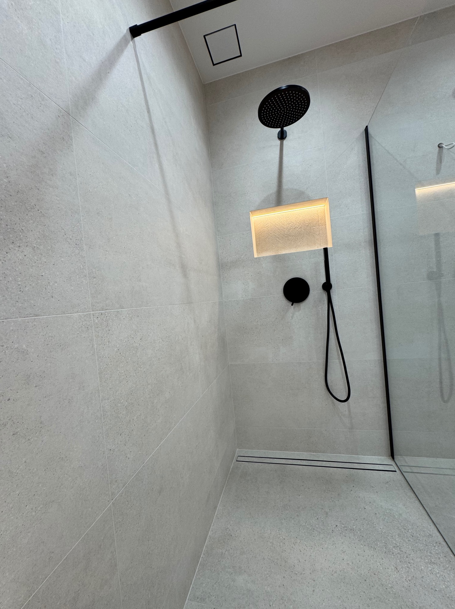 Nowoczesna kabina prysznicowa z szarymi płytkami, czarną armaturą i podświetlaną wnęką. Minimalistyczny design z odpływem liniowym i szklaną ścianką.