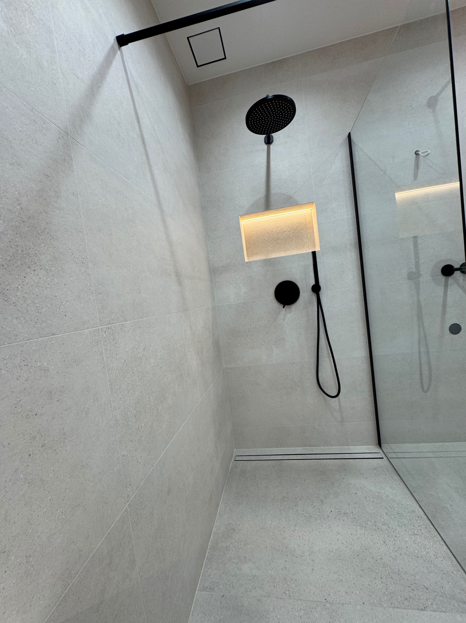 Nowoczesna kabina prysznicowa walk-in z szarymi płytkami, czarną armaturą i podświetlanym panelem. Minimalistyczny design z odpływem liniowym.