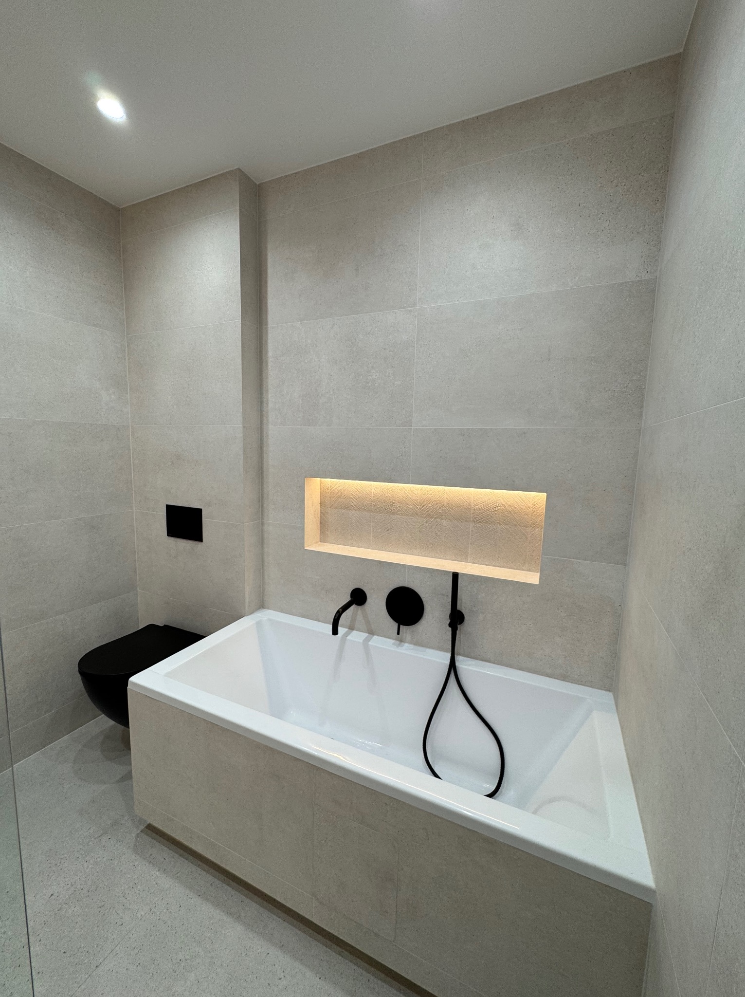 Minimalistyczna łazienka z beżowymi płytkami, czarną armaturą i toaletą. Wnęka nad wanną z subtelnym oświetleniem. Nowoczesny design i stonowana kolorystyka.