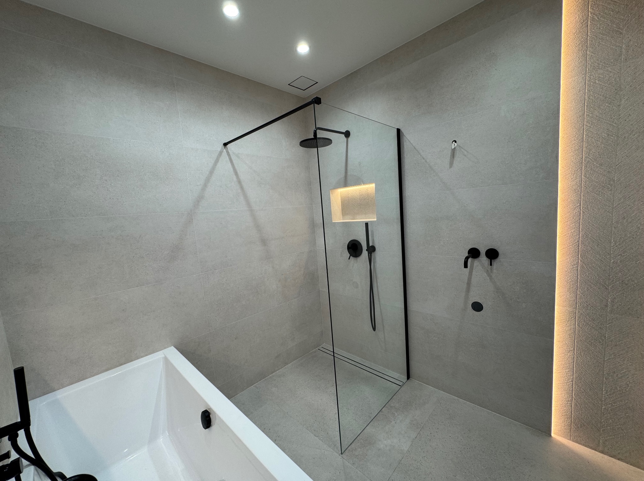 Nowoczesna łazienka z kabiną typu walk-in, czarną armaturą i subtelnym oświetleniem LED. Minimalistyczny design, jasne płytki i wanna.