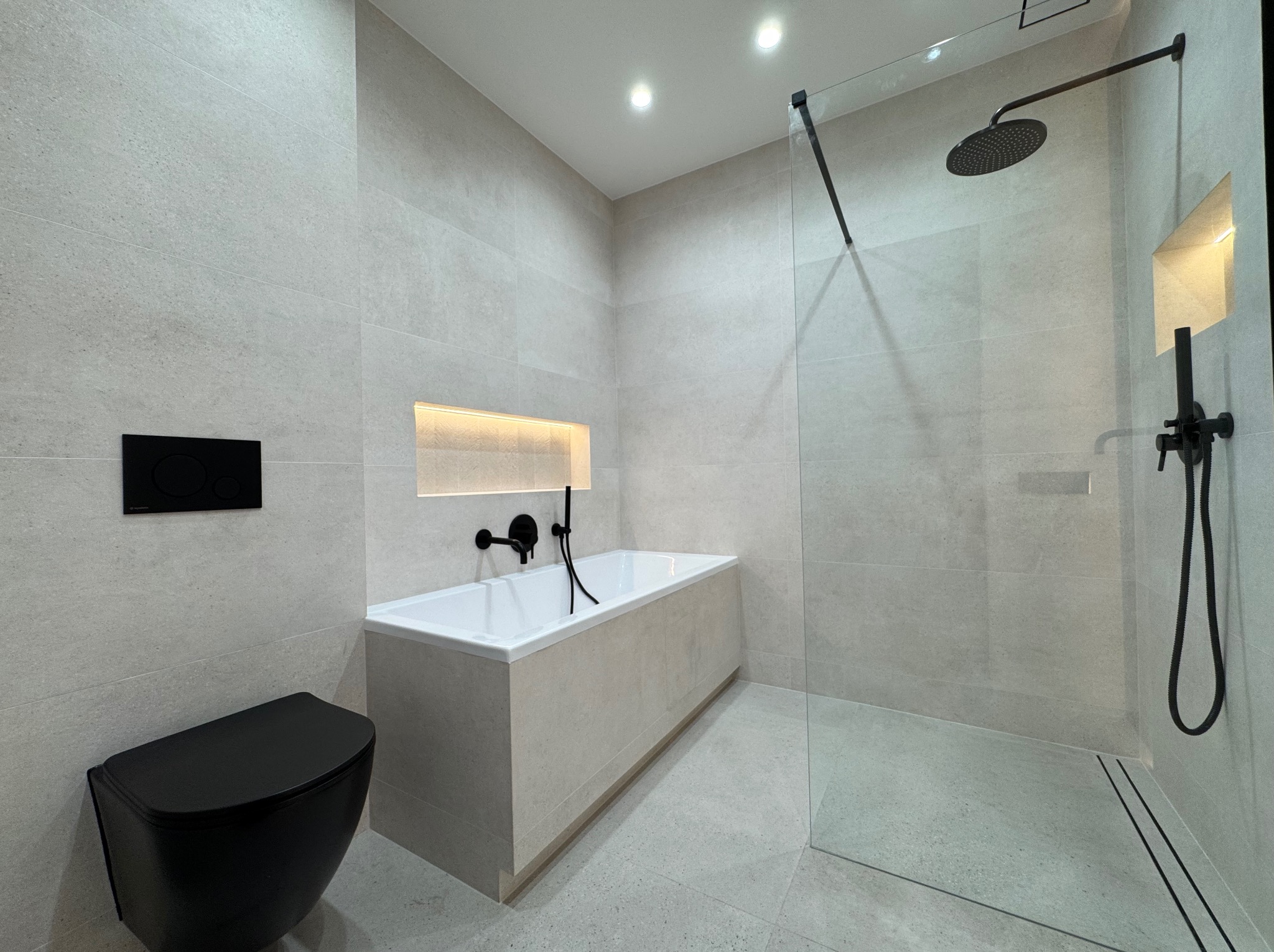 Nowoczesna łazienka w jasnych odcieniach szarości z czarną armaturą: toaleta, bateria wannowa i deszczownica za szklaną ścianką. Wnęka z oświetleniem nad wanną.
