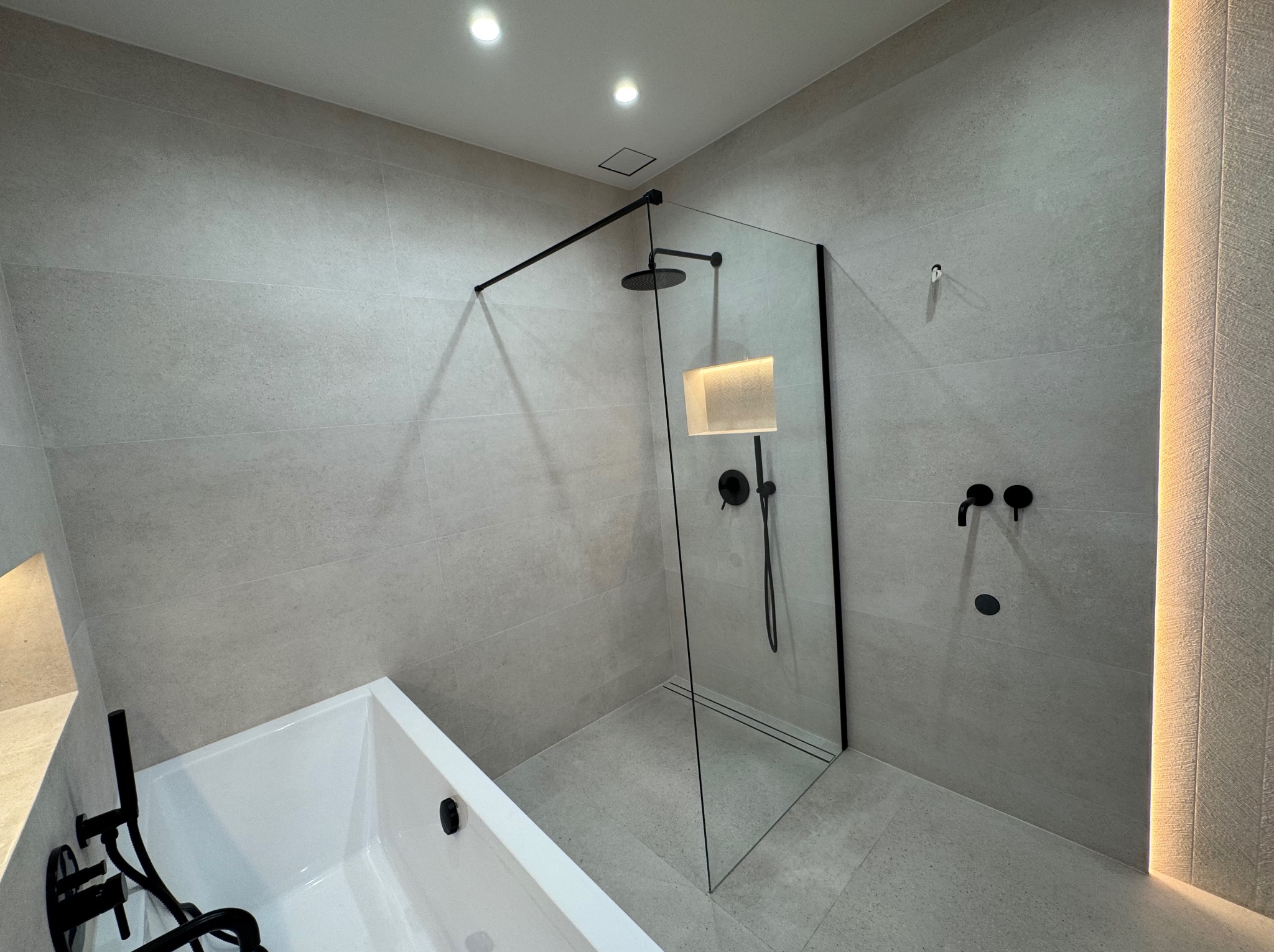 Nowoczesna łazienka z kabiną typu walk-in, białą wanną i czarną armaturą na tle szarych płytek. Oświetlenie LED podkreśla teksturę ściany.