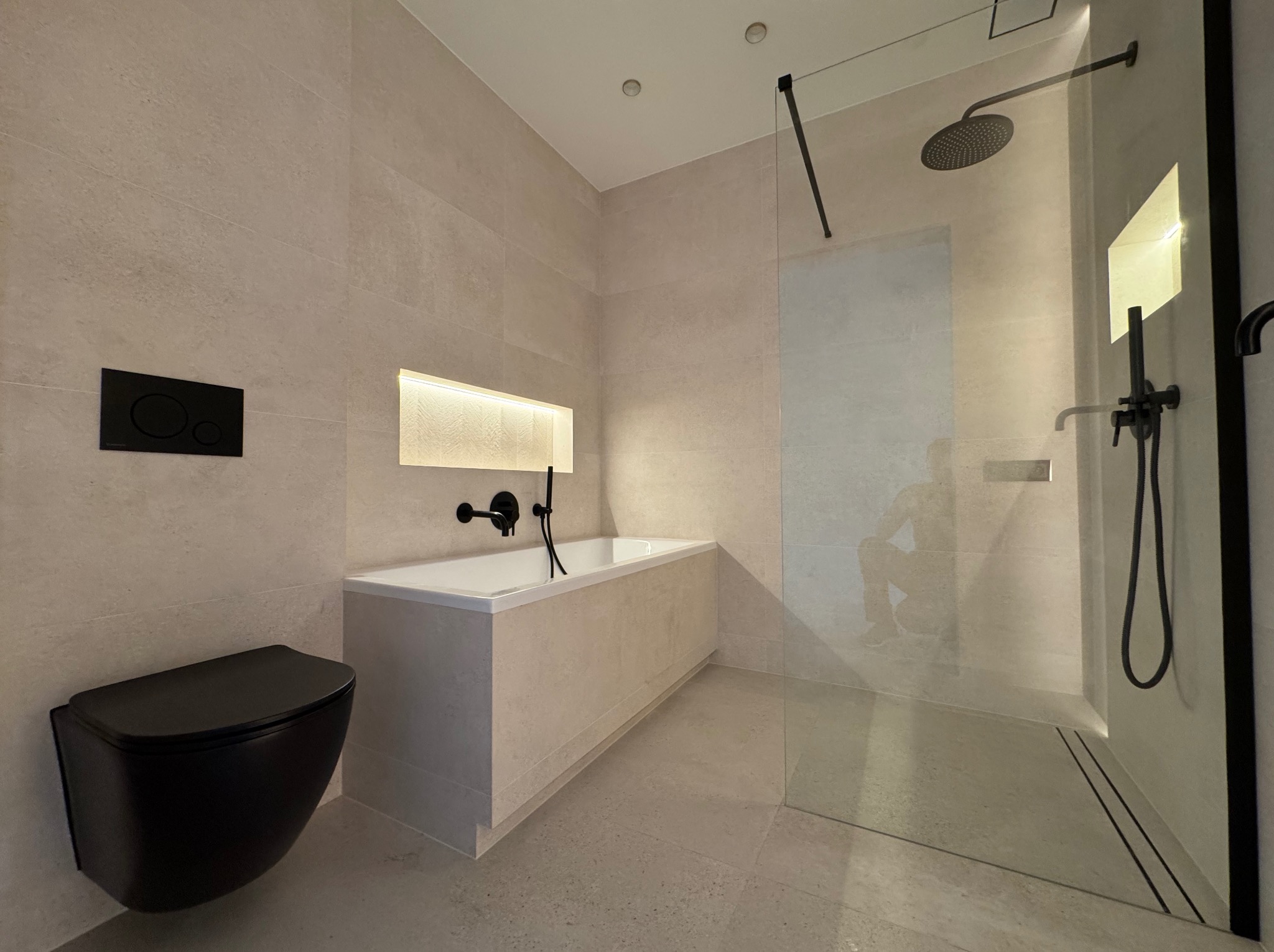 Nowoczesna łazienka w beżowych płytkach z czarną armaturą: bateria wannowa, deszczownica i wisząca toaleta. Wnęka z oświetleniem nad wanną.