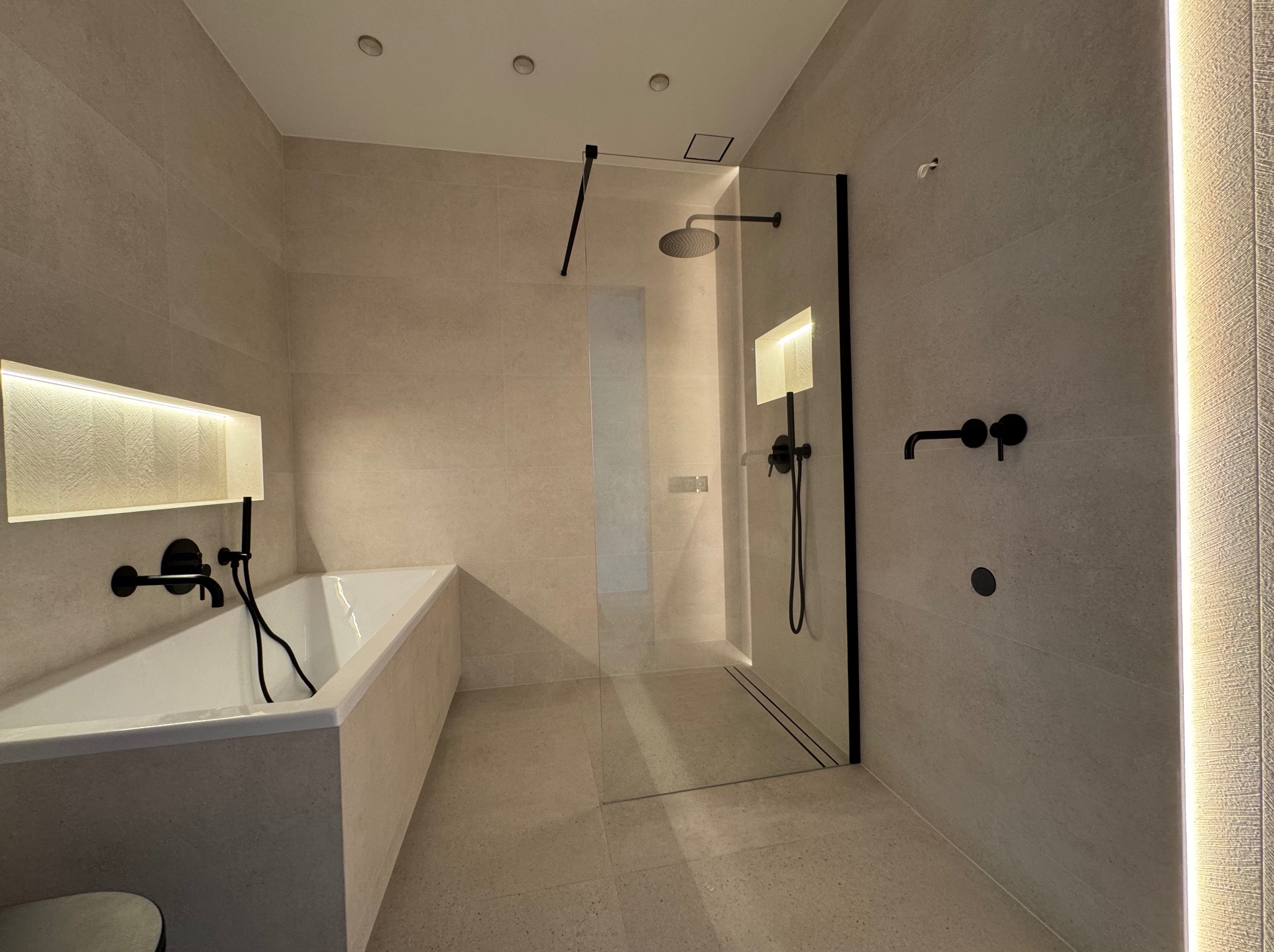 Nowoczesna łazienka w beżowych płytkach z czarną armaturą: wanna z podświetleniem, kabina prysznicowa typu walk-in z odpływem liniowym i oświetlenie dekoracyjne.