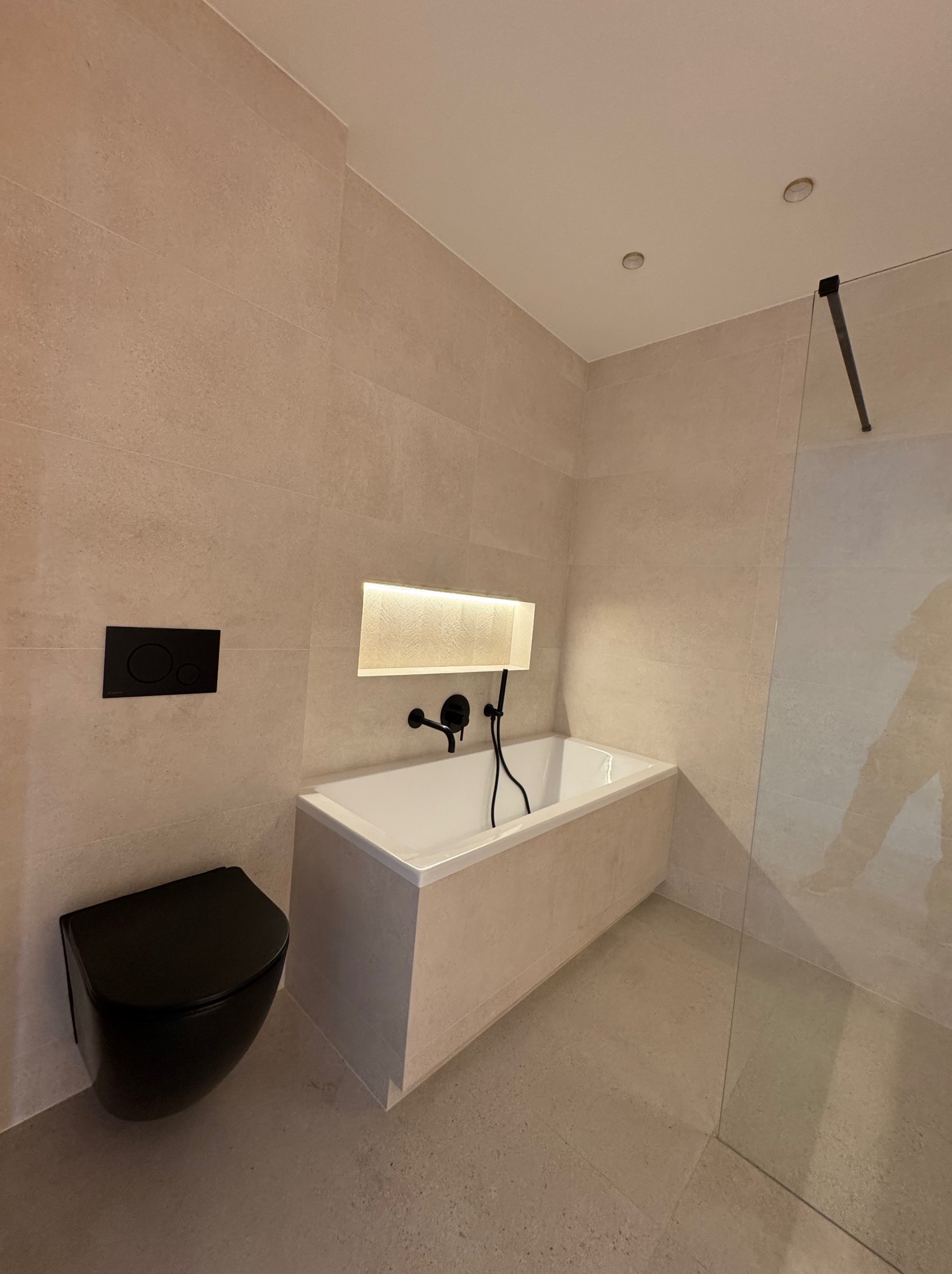 Nowoczesna łazienka w beżach z czarną armaturą: wisząca toaleta, wanna z podświetleniem wnęki, szklany panel prysznicowy. Minimalistyczny design, eleganckie wykończenie.