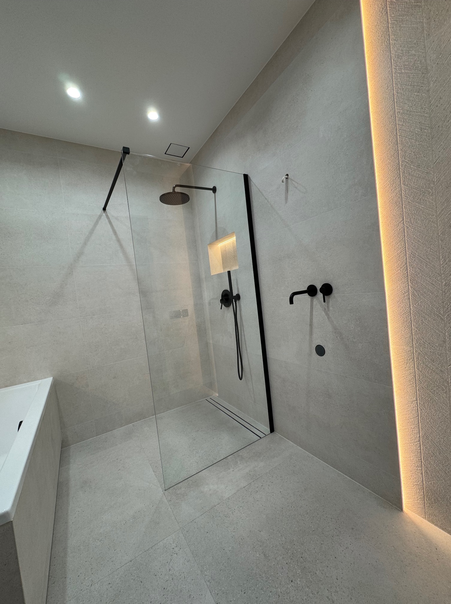 Nowoczesna łazienka z kabiną typu walk-in, czarną armaturą i podświetlanym panelem ściennym. Minimalistyczny design, jasne płytki, eleganckie wykończenie.