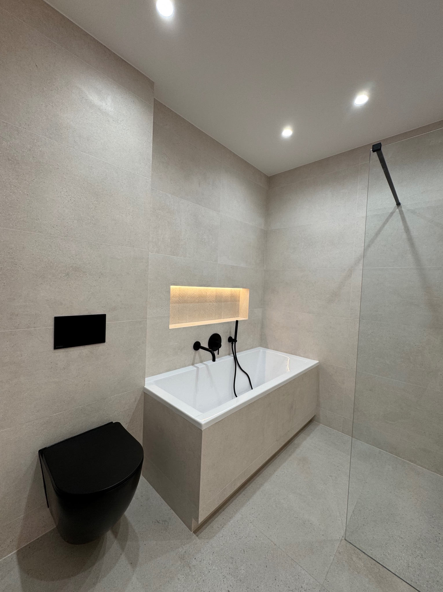 Nowoczesna łazienka w beżowych płytkach z czarną armaturą: wisząca toaleta, bateria wannowa i szklana kabina. Oświetlenie punktowe i wnęka z podświetleniem.